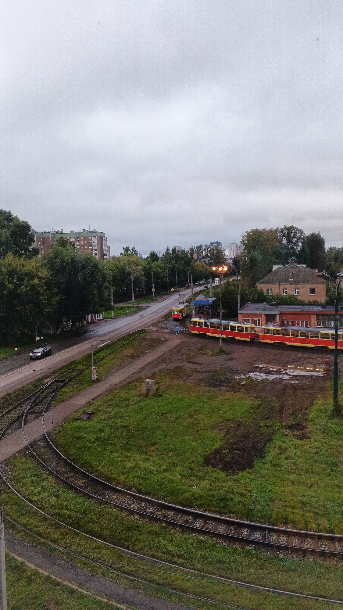 The image depicts an overcast day in Izhevsk, Russia, with a view of a street and tram tracks. In the foreground, several sets of tram tracks curve through a grassy area, some appearing disused. Beyond the tracks, a road is visible, wet with what appears to be rain. A dark-colored car is driving on the wet road. To the right of the road, a tram depot or station is present, with several red and yellow trams parked. One tram is stopped at the platform, and another is parked further back. A person in a blue shirt is walking near the platform. In the background, apartment buildings and trees line the street, with a cluster of taller buildings visible in the distance. The sky is uniformly grey with clouds.