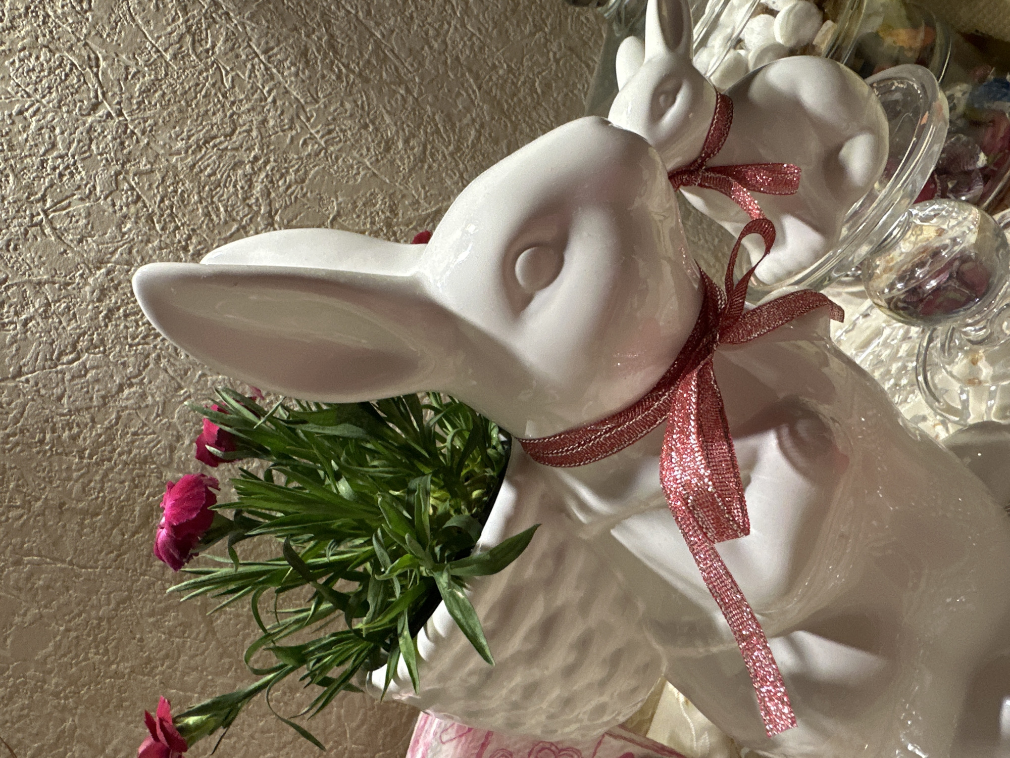 A close-up shot shows two glossy white ceramic rabbit figures with red ribbons around their necks. The larger rabbit holds a bouquet of green stems and small pink carnations. The background includes textured wallpaper and a glass bowl filled with white candies and other small decorative items. The image appears to be taken indoors, with lighting that casts shadows and highlights the reflective surfaces. No people are visible. The location is not discernible from the image.