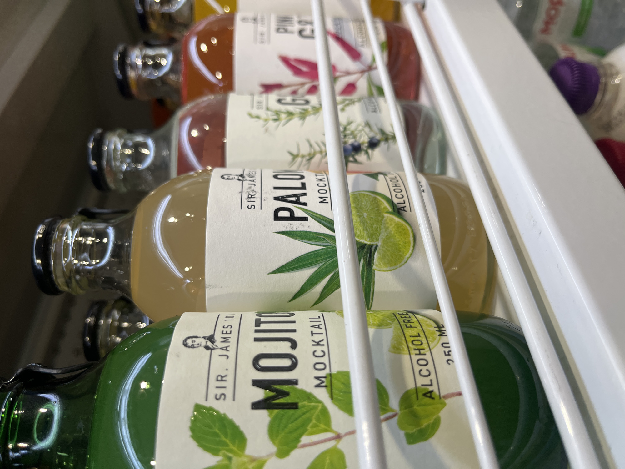 The image shows a row of bottled beverages on a white shelf, likely in a store or on display. The bottles are arranged horizontally, and the focus is on the labels of the drinks. The first bottle on the left is dark green and has a label that reads "MOJITO MOCKTAIL" and "ALCOHOL FREE 250 ML" with an image of mint leaves. Next to it is a bottle with a light brown liquid and a label that says "PALOMOCKTAIL." Further down the shelf, other bottles are visible, some with red or pink liquids, and labels that partially read "GO" and "PINK CRUSH." The bottles appear to be part of a product line from "SIR. JAMES 101." The lighting suggests an indoor environment. The background is dark and out of focus.