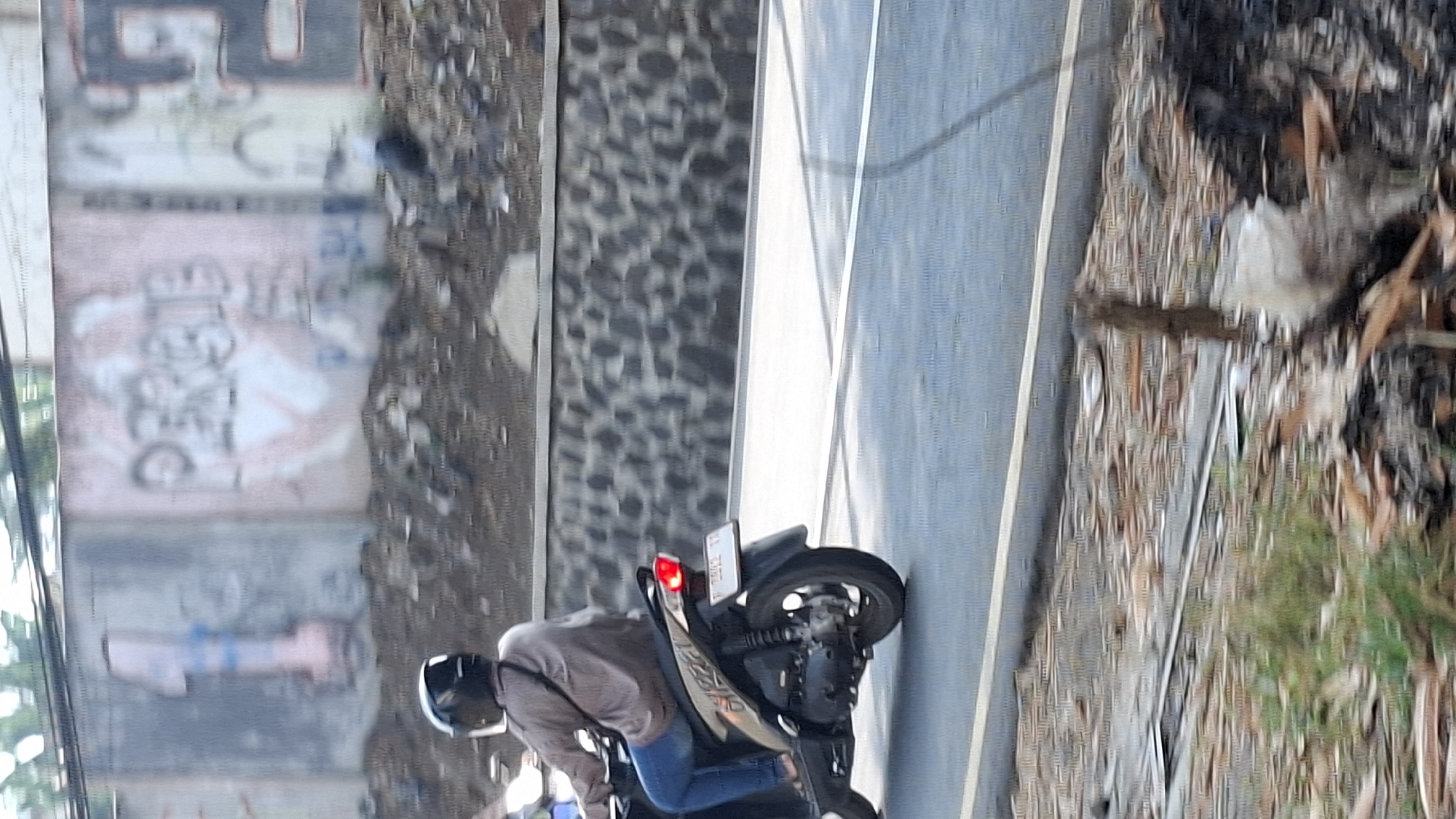 A person wearing a black helmet and a brown long-sleeved shirt rides a dark-colored scooter along an asphalt road. The scooter has a license plate with white characters on a black background. The road is bordered by a low stone wall on the left, with graffiti on the wall behind it. The ground in front of the wall is covered with debris. Trees and sky are visible above the wall.