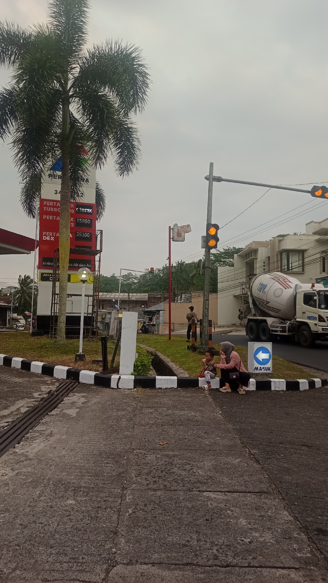 The image depicts an outdoor scene, likely in the city of Karyamandala, Indonesia, with a focus on a street intersection. In the foreground, a concrete road with black and white striped curbing is visible, along with a metal grate drain. To the right, a woman and a young child are seated on the curb. The woman is wearing a pink hijab and a pink long-sleeved shirt with dark pants. The child is wearing a grey and brown patterned long-sleeved shirt and grey pants. They appear to be interacting.
Behind them, a large concrete mixer truck with "TIMAX" branding on its side is parked on the road. A traffic light with amber signals is illuminated. A sign with a blue arrow and the word "MASUK" (meaning "enter" in Indonesian) is posted nearby.
To the left of the scene, a tall palm tree stands next to a gas station pylon displaying prices for various fuel types, including "PERTA X TURBO" and "PERTA DEX". A red awning of the gas station is partially visible. The background features buildings, trees, and an overcast sky. A man is standing near a post further back on the right side of the road, and several other individuals and market stalls are visible in the mid-ground. The overall atmosphere suggests a typical day in a semi-urban environment.