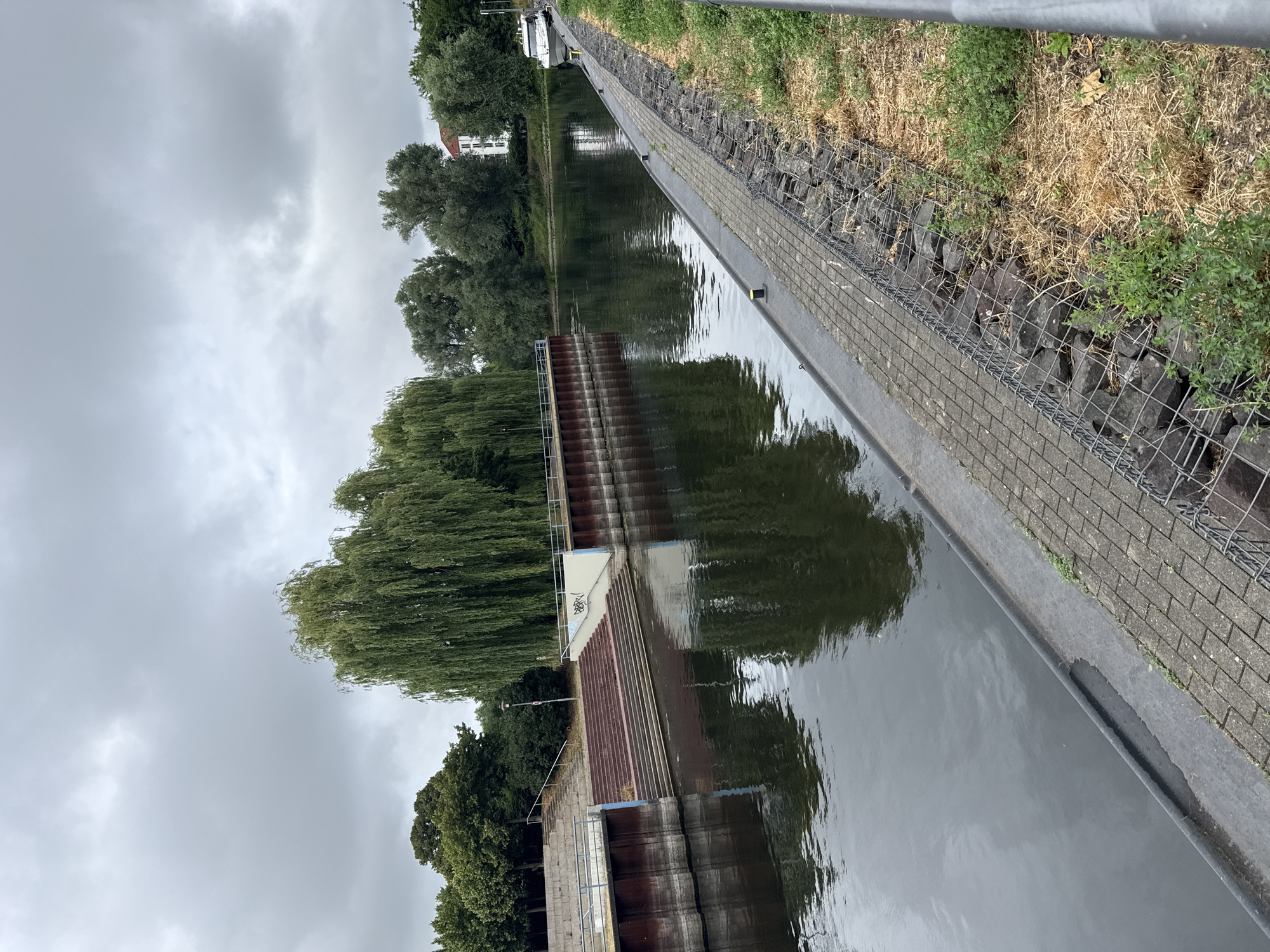 A serene canal scene unfolds in Rathenow, Germany, under a heavy, overcast sky. The water, dark and reflective, mirrors the lush greenery and architecture lining its banks. On the left, a wide, tiered concrete staircase descends to the water's edge, bordered by a modern, dark metal retaining wall with horizontal lines. Beyond the stairs, a cluster of trees, including a prominent weeping willow with its cascading branches, creates a dense green backdrop.
The canal itself stretches into the distance, appearing calm and unruffled. On the right bank, a paved walkway runs alongside the water, constructed with interlocking gray bricks. This walkway is bordered by a gabion wall filled with large stones and wire mesh, separating it from a strip of dry grass and scattered green vegetation. Further along the right bank, a white motorboat is docked, its sleek form a contrast to the natural elements. In the background, a white building with red-tiled roofs stands amongst more trees, hinting at the presence of urban life beyond the tranquil waterway. The overall atmosphere is one of quietude and natural beauty, punctuated by man-made structures.