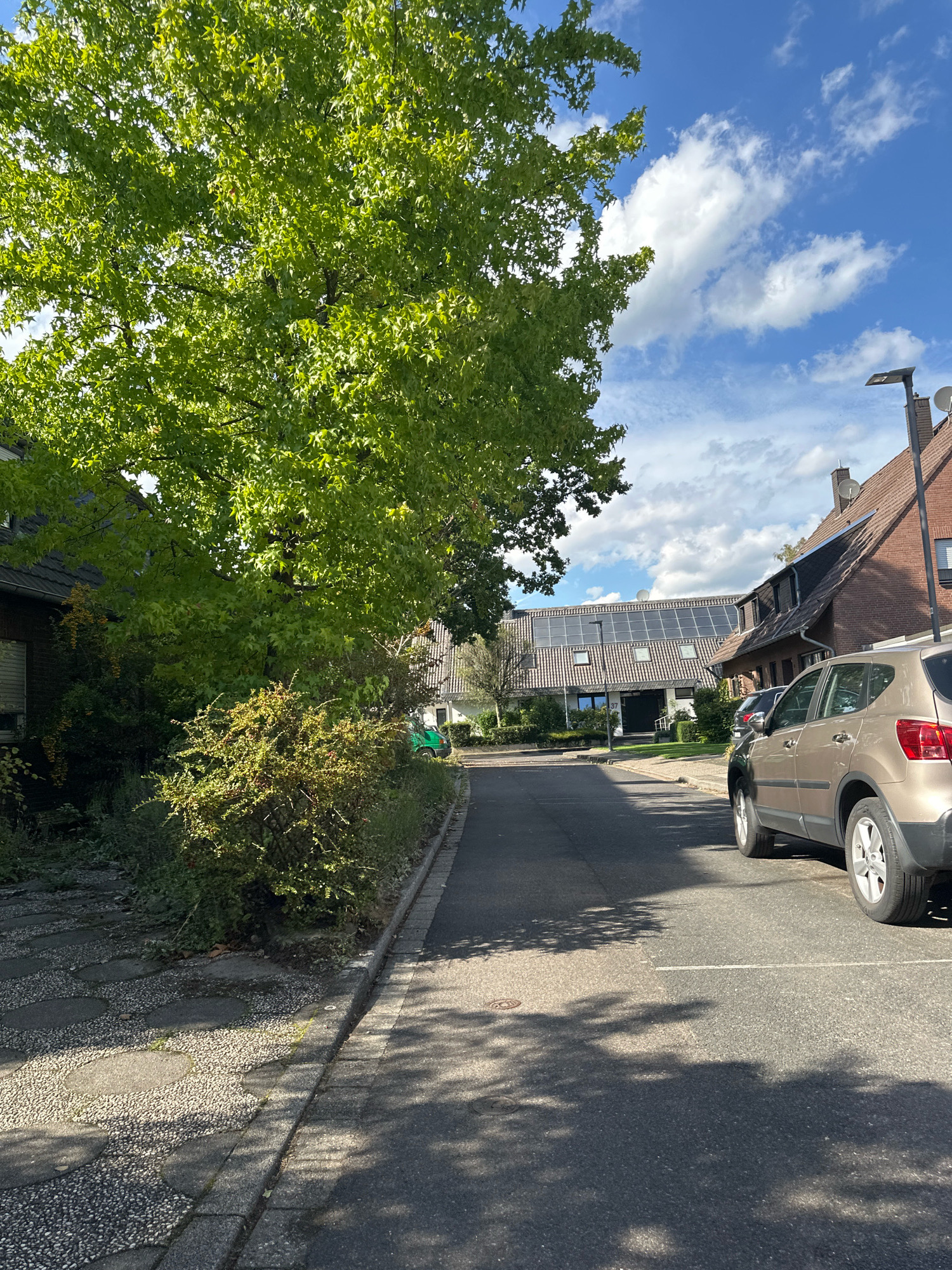 The image depicts a street in Krefeld, Germany, on a sunny day with scattered clouds. On the left side of the street, there is a large tree with vibrant green leaves casting shadows on the pavement. Adjacent to the tree is a pathway made of gravel and circular stone pavers. Further down the street, several residential buildings are visible, some equipped with solar panels. A light-colored car is parked on the right side of the street, facing away from the camera. A green vehicle is parked further down the street on the left. The overall scene is residential and appears quiet.