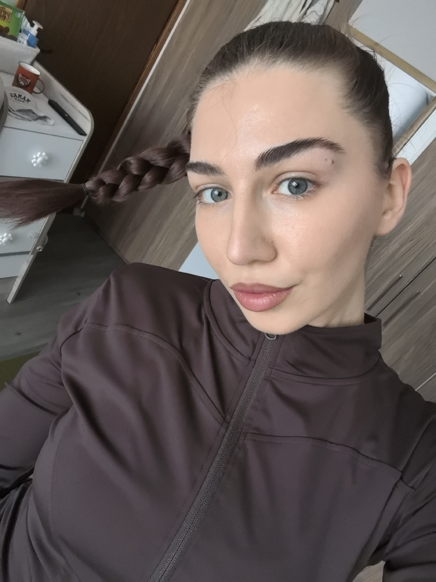 A medium shot photograph captures a woman with fair skin and light eyes, likely indoors. Her dark hair is pulled back into a braid that extends to the left side of the frame, close to a white dresser. The woman wears a dark brown jacket with a zipper. The background includes wooden furniture and a partially visible doorway. The location in Stara Zagora, Bulgaria, is not explicitly ascertainable from the visual data.