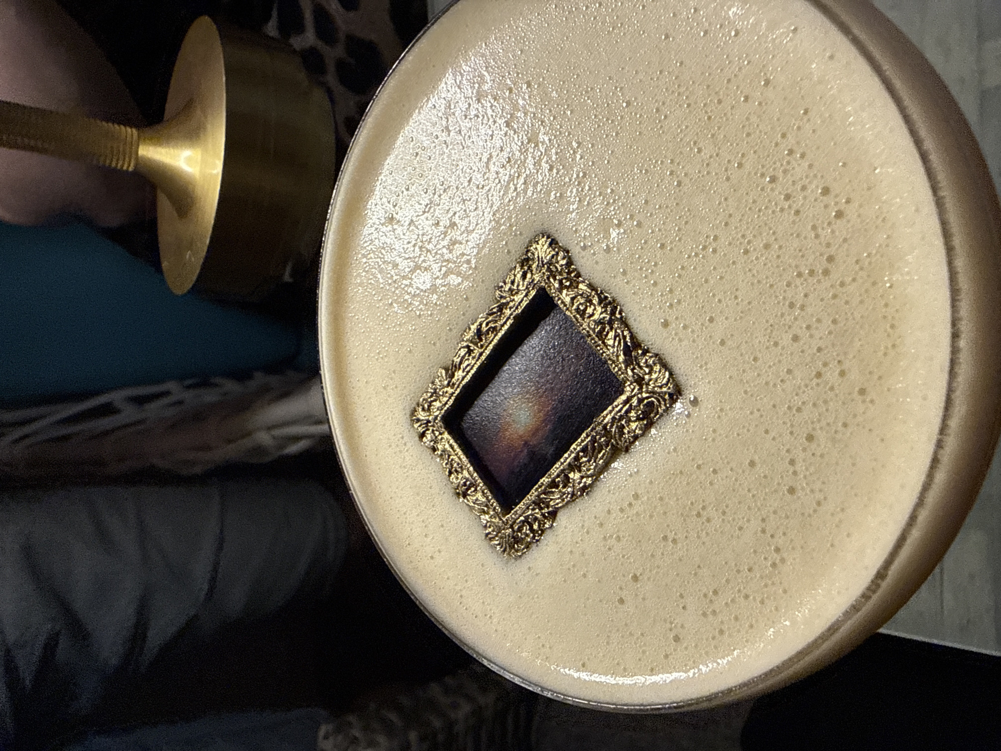 A close-up, top-down view captures a creamy, light-brown foamy drink in a glass. Floating in the center of the foam is an ornate, gold-colored diamond-shaped frame. Inside the frame, a dark, iridescent surface shimmers with hues of purple, blue, and orange, suggesting a liquid or solid inlay. The foam around the frame is dotted with tiny bubbles, indicating it is freshly poured. The background is out of focus, showing dark fabric and a glimpse of a gold lamp with a flared base. No individuals are clearly visible. The location in Feodosia, Ukraine, is not discernible from the image.