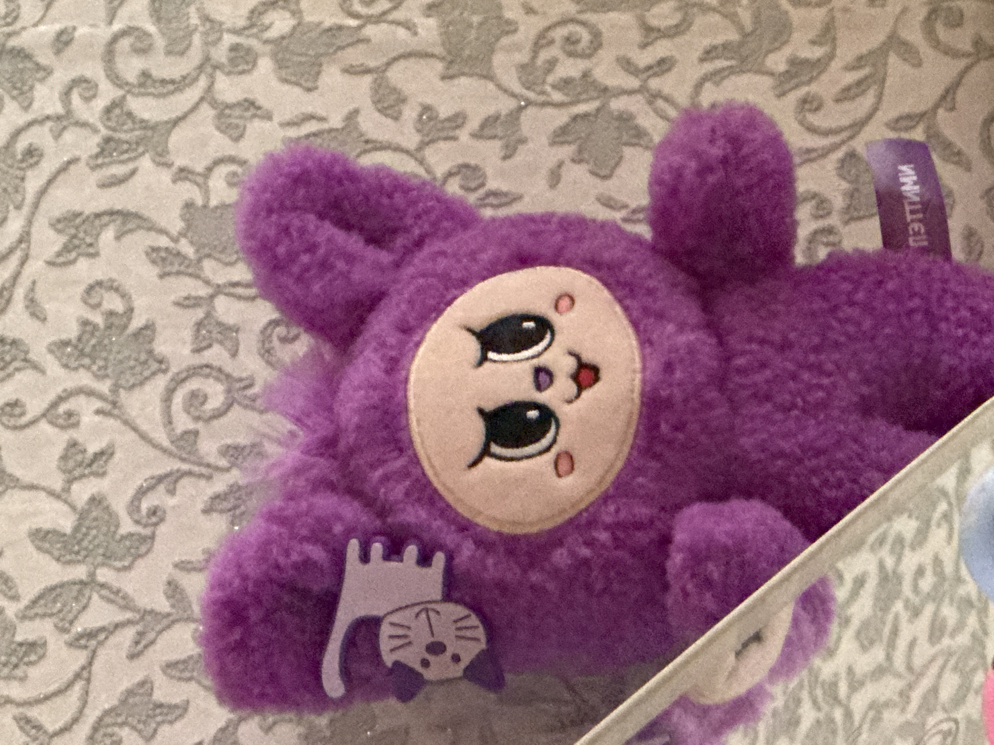 A close-up, eye-level, indoor shot shows a purple, plush toy with large, dark eyes, a small pink mouth, and rosy cheeks. The toy is positioned in the center of the frame against a grey and white patterned background. A small, light purple accessory featuring a stylized cat face is attached to the toy's left ear. A purple tag with white Cyrillic text is visible on the lower right side of the toy. The image appears to have been taken in Bratsk, Russia, based on the tag text.