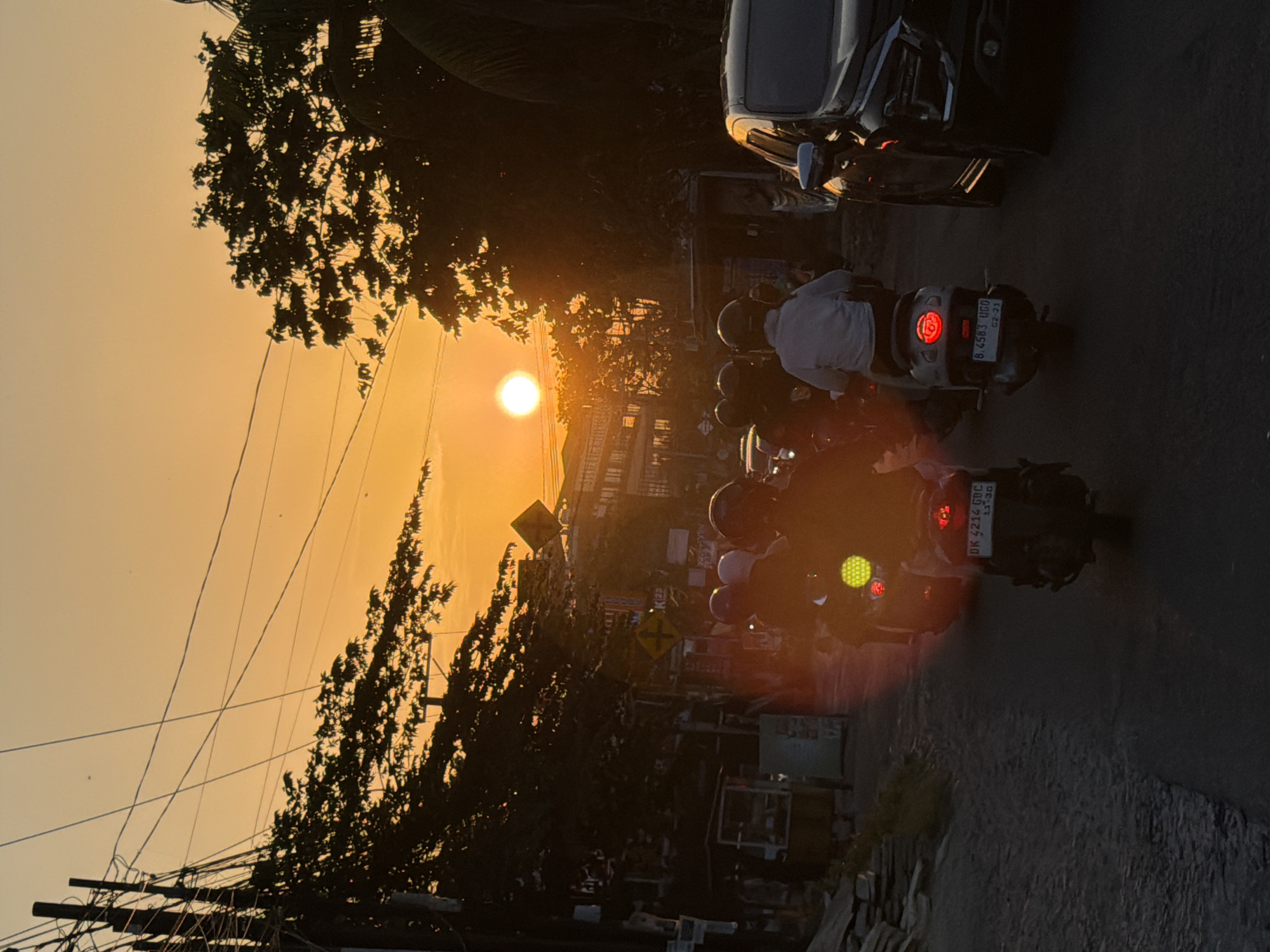 The image depicts a street scene in Mengwi, Indonesia, during a sunset. The sun, a bright orb, is low on the horizon, casting a warm, orange glow across the sky and silhouetting trees and utility poles. A paved road runs through the scene, with several motorcycles in motion, their headlights and taillights illuminated. The riders are wearing helmets. A dark-colored SUV is parked on the right side of the road. Buildings line the left side of the street, some appearing to be shops or residences. The overall atmosphere is that of a busy street at dusk.