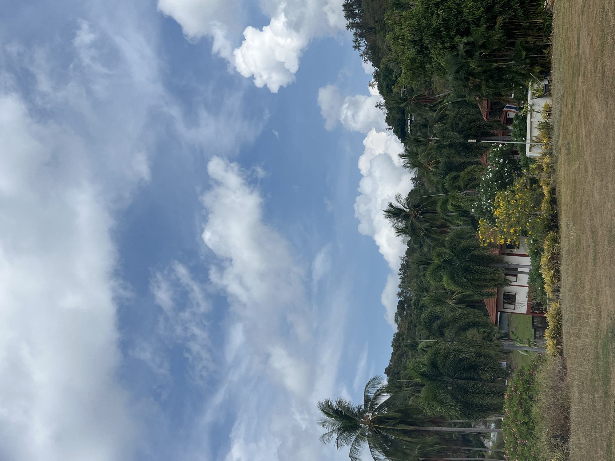 The image shows a partially cloudy sky above a tropical landscape in Baan Hua Thanon, Thailand. A hillside covered with dense green trees and palm trees is visible on the right side of the frame. A building with a red roof is visible among the trees, and a white structure with a sign that says "LABWAN VIEW POINT" can be seen as well. A grassy area is visible in the foreground.