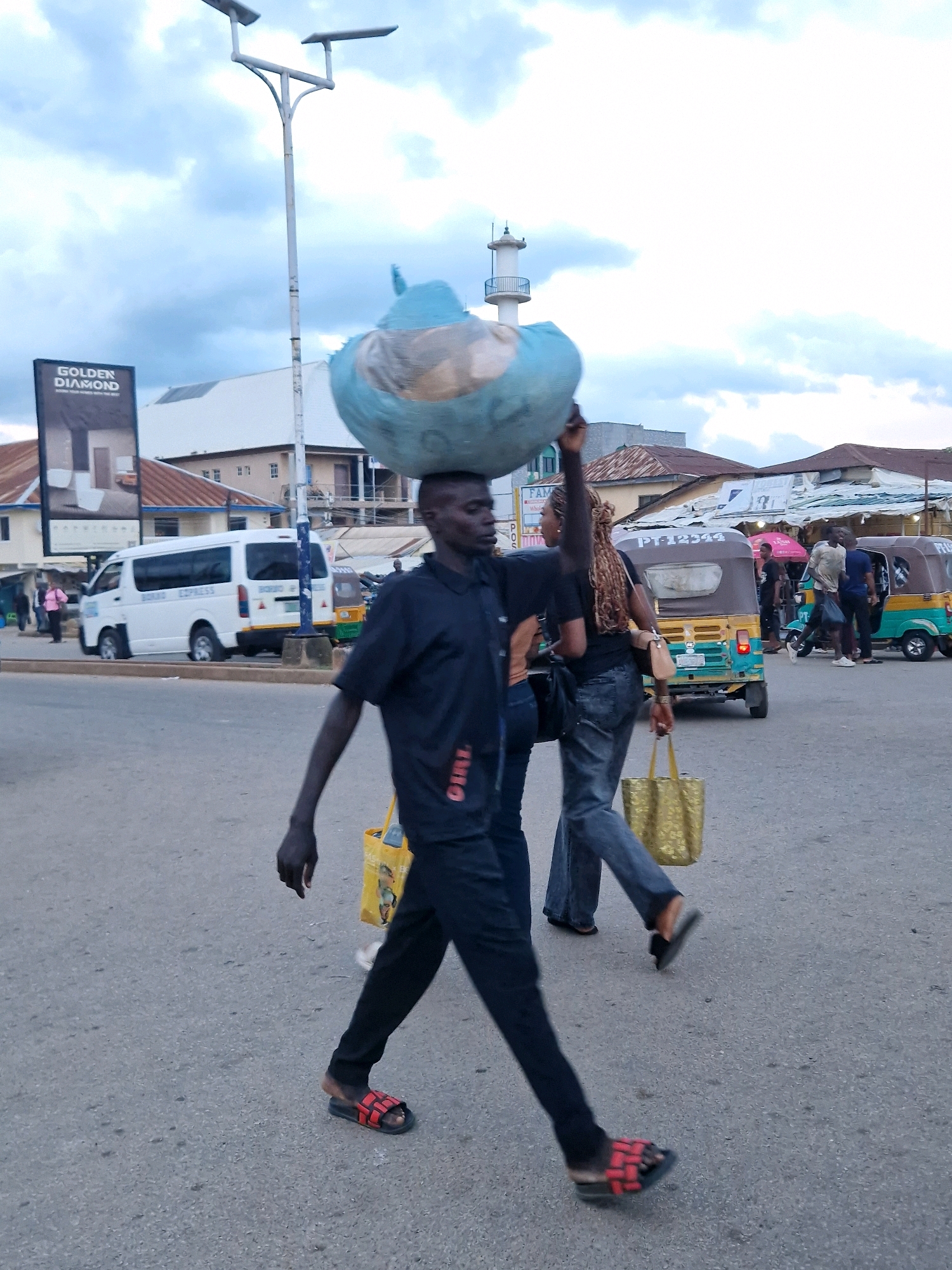 A man walks across a paved street in Jos, Nigeria, balancing a large, light blue object wrapped in plastic on his head. He is wearing a dark blue short-sleeved shirt and dark pants, with red and black slide sandals on his feet. A yellow shopping bag is slung over his left shoulder. Behind him, a woman with braided hair walks with her right arm raised, also carrying a gold tote bag. In the background, a white van with "BOMU EXPRESS" written on it is parked on the side of the road. Other vehicles, including a yellow and green auto-rickshaw, and various buildings and market stalls are visible. The sky is overcast.