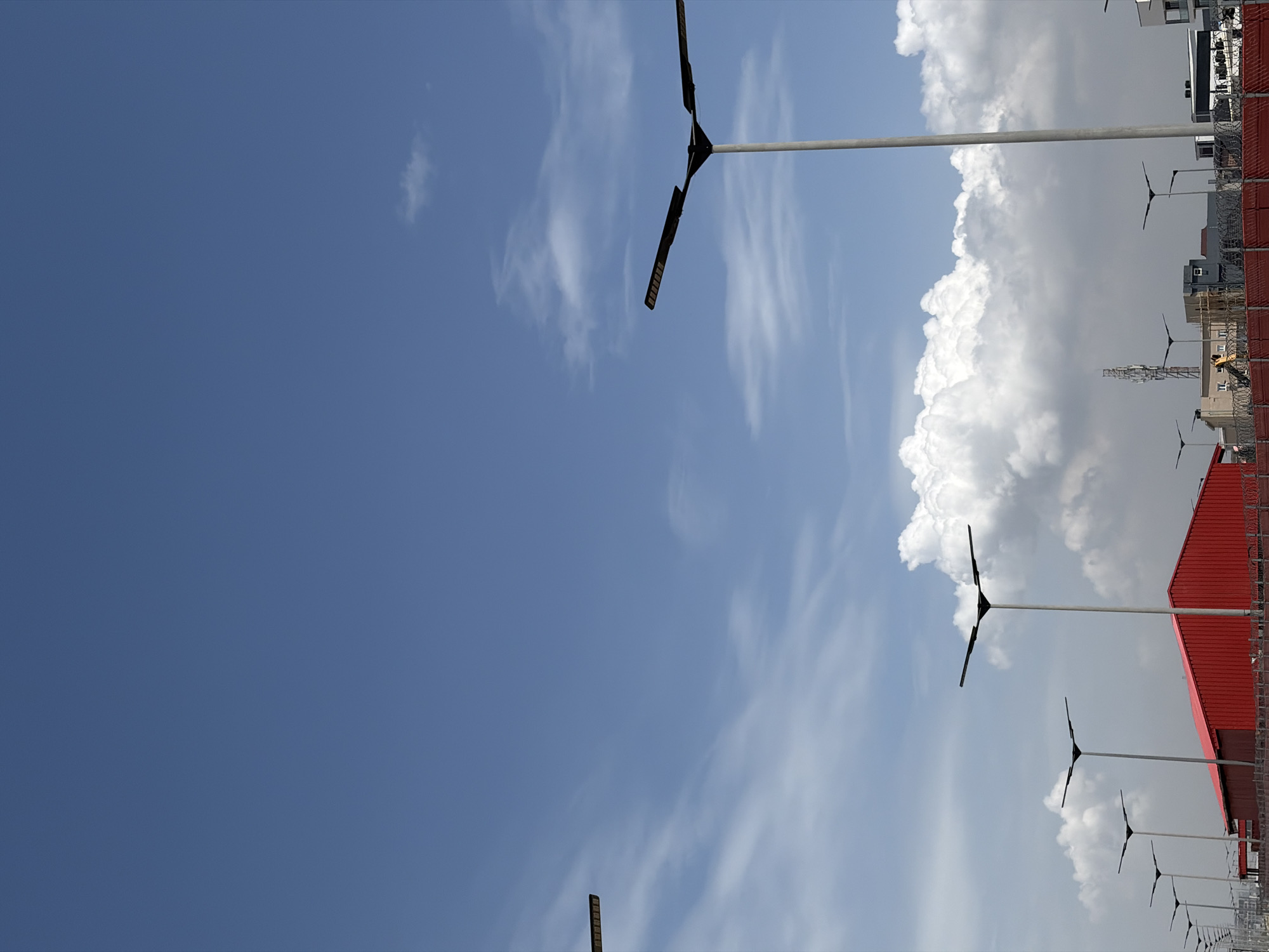 The image shows a view of the sky above Igboefon, Nigeria. A gradient of blue sky transitions into an area with white cumulus clouds. Several tall streetlights with a distinctive "T" shaped head are lined up along the bottom edge of the frame. The tops of buildings and a red roof are also visible.