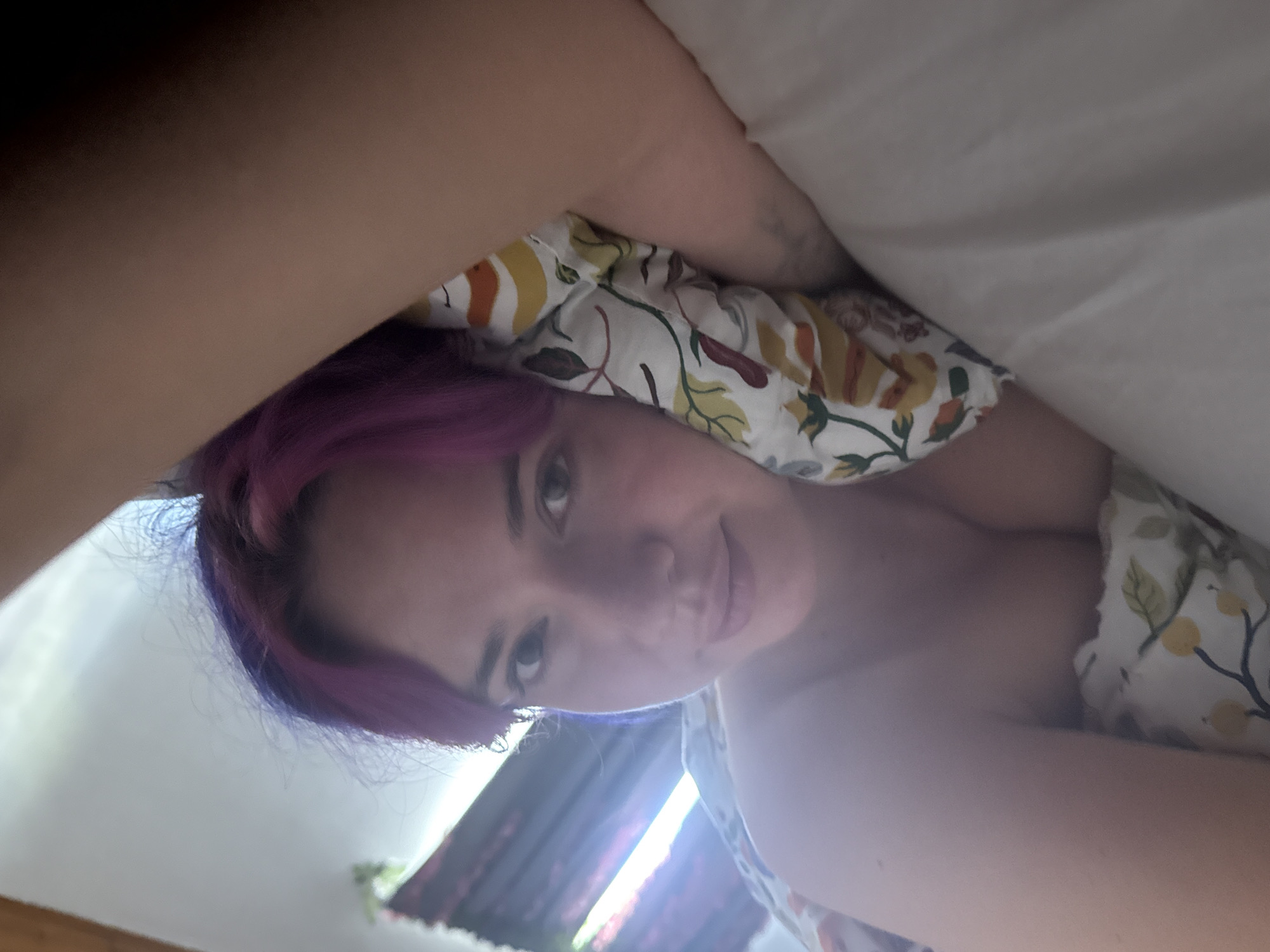 A young woman is captured in a close-up, angled shot, likely taken from a low perspective while she is lying in bed. Her head is positioned towards the center-right of the frame, with her body partially obscured by bedding and a bare arm in the foreground.
The woman has distinctive purple hair, with some darker roots visible, and her hair is somewhat disheveled, suggesting she might have just woken up or is relaxing. She has light-colored eyes and a soft, slight smile on her lips, conveying a relaxed and pleasant emotion. Her gaze is directed towards the camera.
She is partly covered by bedding, which includes a white sheet and a pillow or duvet with a patterned fabric. This fabric features a design of autumn-like leaves and berries in shades of yellow, orange, and green on a white background. A bare shoulder and arm are visible, suggesting she might be wearing a simple top or be partially covered by the sheets.
In the background, a bright light source, likely a window, is visible, creating a strong backlight and some lens flare around her head and hair. This indicates it is daytime. The overall setting appears to be an indoor bedroom in the City of Zagreb, Croatia, providing a personal and intimate atmosphere. No visible text is present in the image.