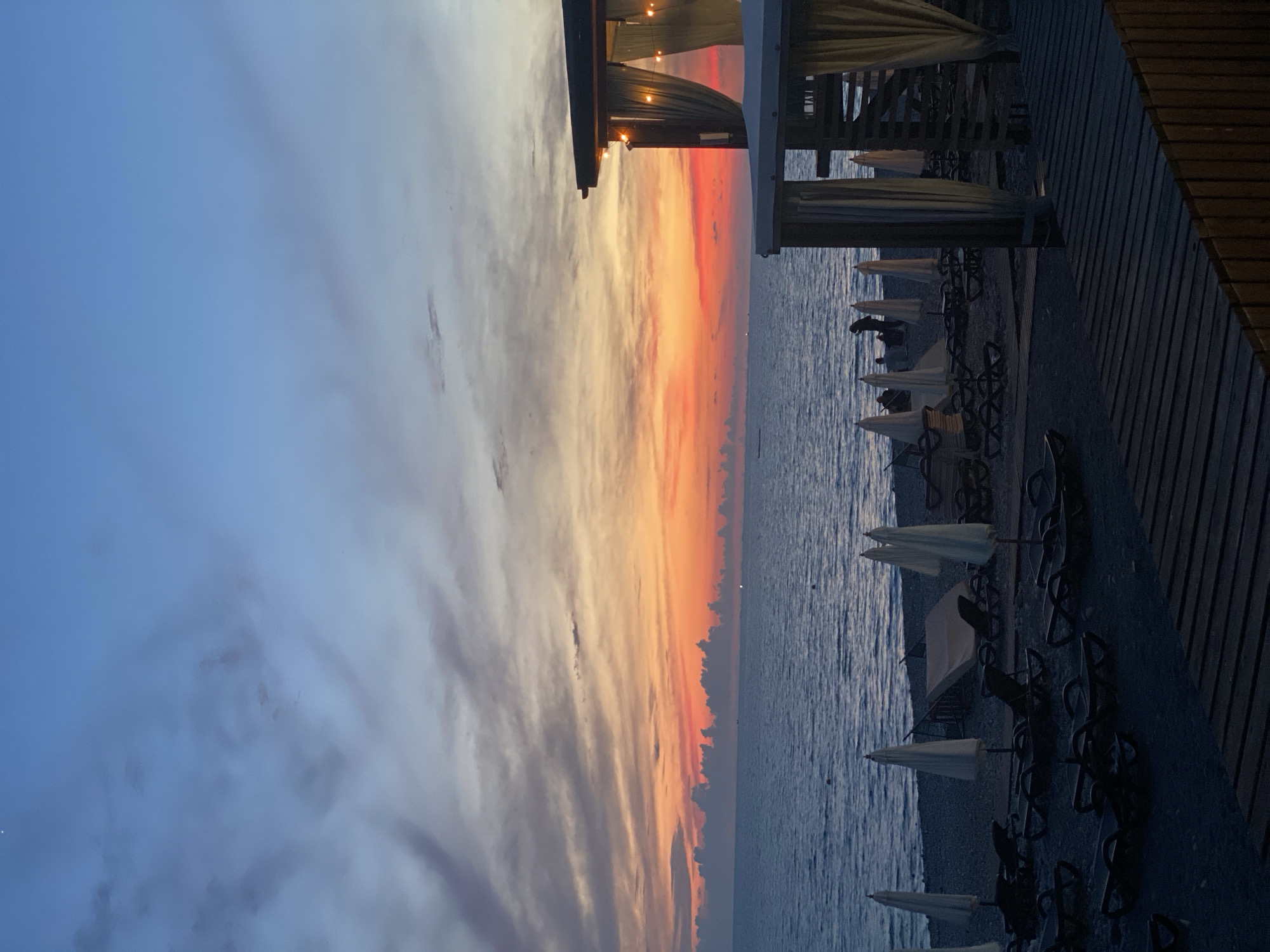 The image depicts a serene beach scene in Sochi, Russia, during twilight. The sky is a canvas of vibrant colors, transitioning from deep blues at the top to fiery oranges and reds near the horizon, with scattered clouds adding texture and depth. The calm sea stretches out to meet the colorful sky, with a few distant lights visible on the water, possibly from boats or buoys.
On the shore, rows of empty lounge chairs and closed umbrellas are neatly arranged on the pebbled beach, suggesting the end of the day's activities. In the foreground, wooden decking provides a pathway or viewing area. To the right, a cabana-like structure with draped curtains and ambient lighting is partially visible, adding a touch of resort ambiance. A few figures are scattered sparsely on the beach, some appearing to be walking or standing near the water's edge, contributing to the tranquil atmosphere. The overall impression is one of peaceful relaxation as the day gives way to night.