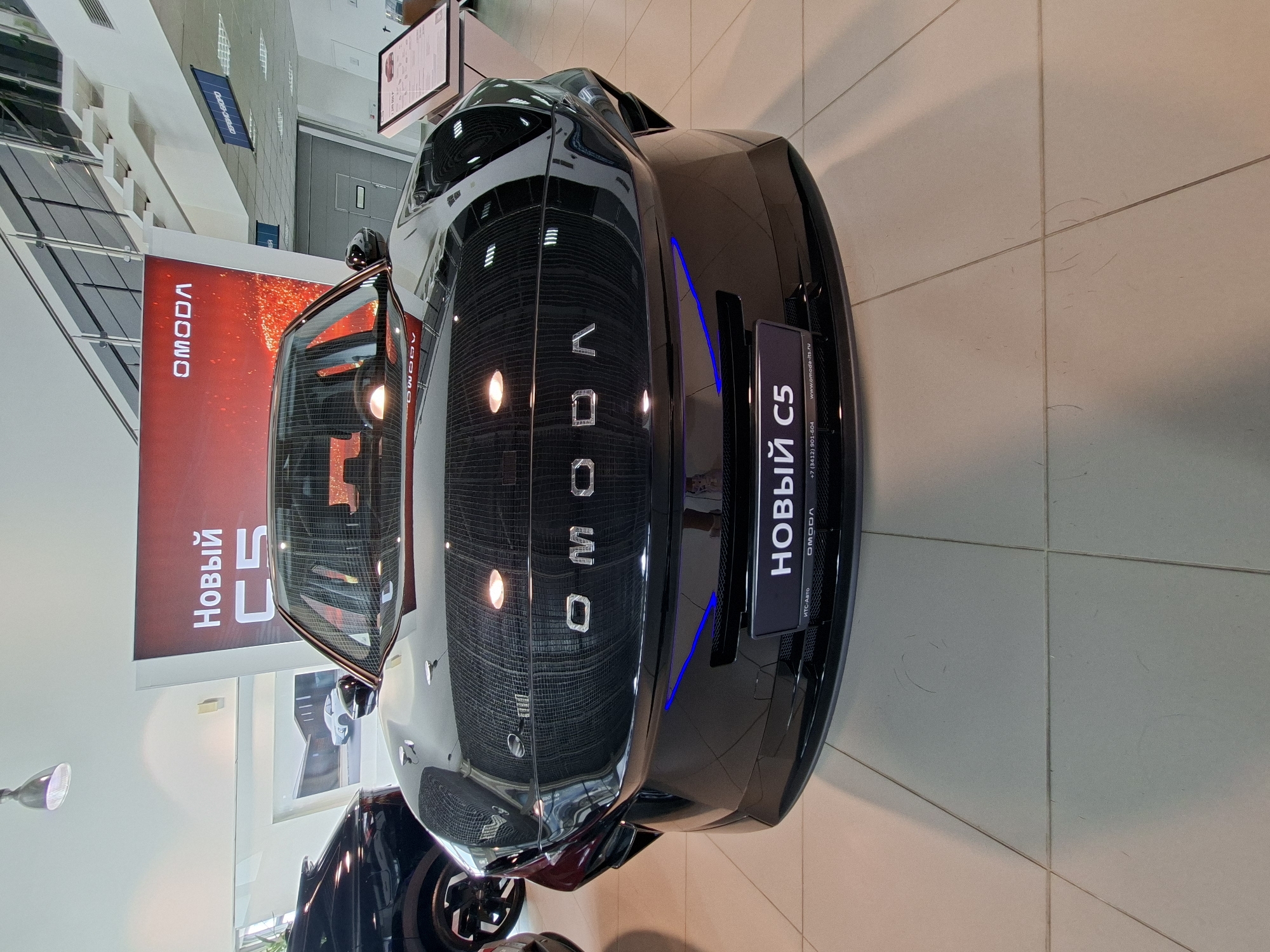 A black OMODA C5 car is parked in a showroom, likely a dealership, in Izhevsk, Russia. The car is the central focus of the image, with its front grille, headlights, and logo clearly visible. The car has a sleek, modern design, and the showroom appears to be well-lit and spacious, with tiled flooring and a large promotional banner in the background displaying the OMODA logo and text in Russian. The banner reads "НОВЫЙ C5," meaning "New C5," suggesting this is a new model being showcased. Other cars and display items are visible in the background, indicating a commercial environment. The overall impression is that of a professional car dealership displaying a new vehicle.