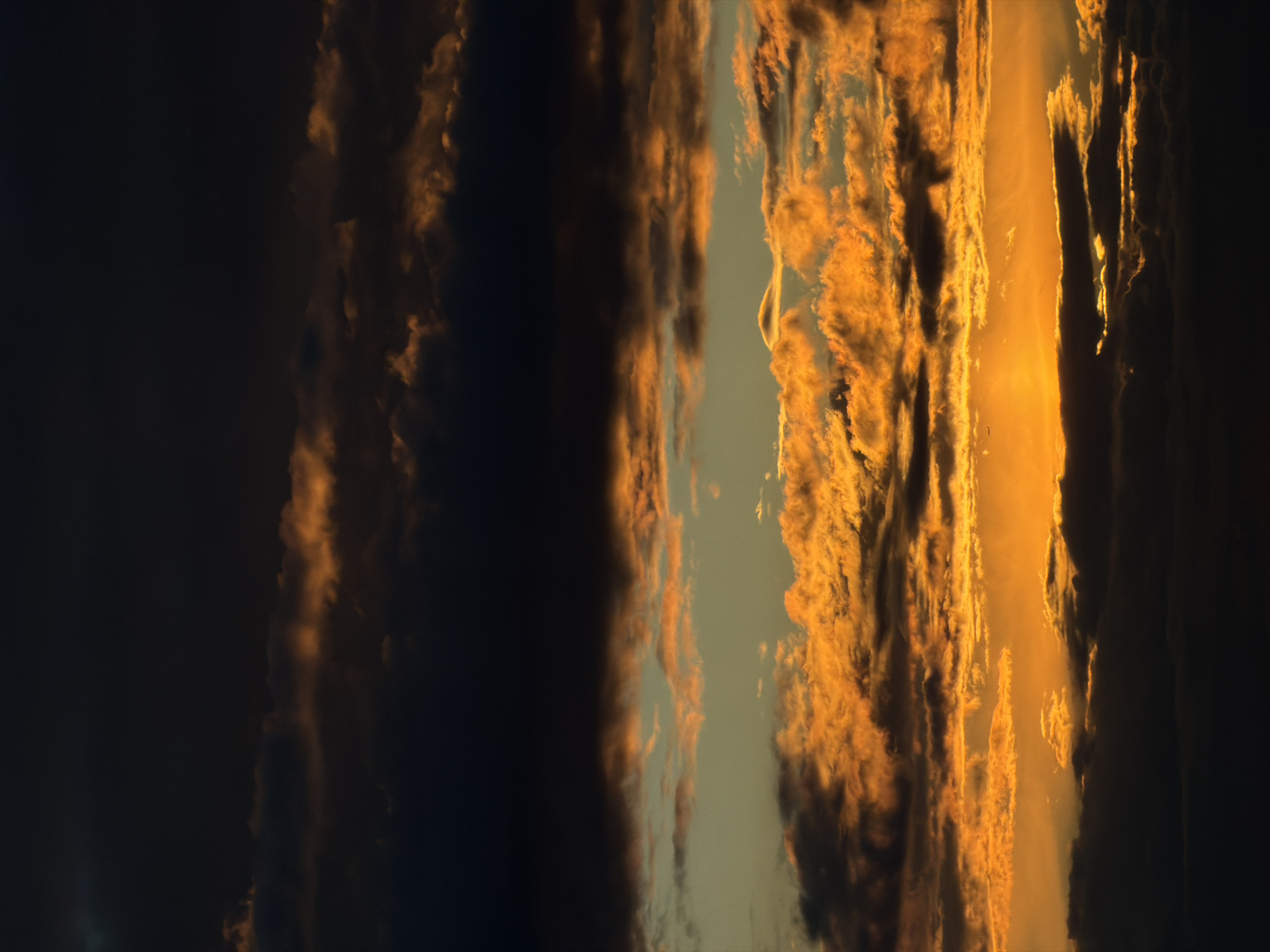The image depicts a vertical view of a sunset with clouds in the sky. The left side of the image is dominated by dark, heavy clouds, while the right side is illuminated with orange and yellow hues, indicating the presence of sunlight. There's a narrow gap between the clouds where the sky is visible. There are no people or specific objects visible. The location is Unknown, Unknown.