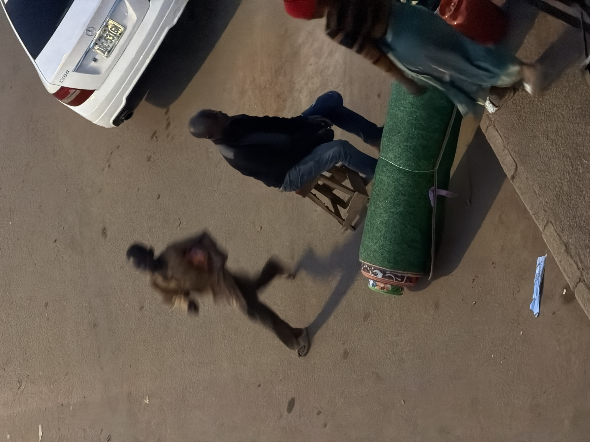 In Bukuru, Nigeria, a blur of motion depicts a person in brown clothing running across a dusty, unpaved street, possibly carrying an object in their arms. To their right, a man in a black jacket and blue jeans sits on a wooden stool, his gaze directed downwards. Further to the right, a person in a light blue skirt and a striped top, with a red head covering, walks away from the camera. In the background, a white Mercedes-Benz sedan is parked on the street. A large, rolled-up green carpet lies on the ground in the foreground, near a red plastic container. The scene appears to be an outdoor, urban environment.