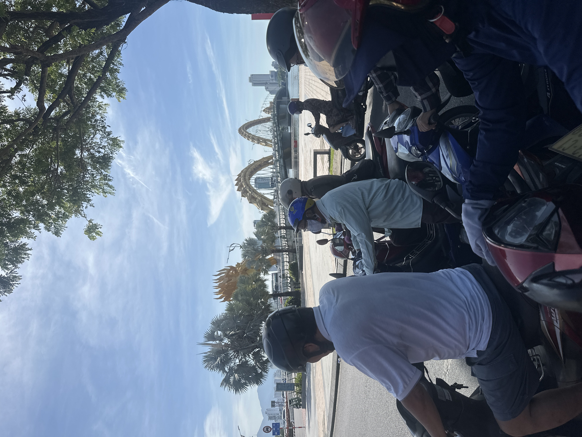 In Đà Nẵng, Vietnam, a group of people on motorbikes waits at an intersection. In the background, the iconic Dragon Bridge spans the Hàn River, its dragon motif visible. The sky is bright blue with wispy white clouds, and a large tree with green foliage provides shade on the right side of the frame. Several palm trees are also visible near the bridge. The people are wearing helmets and casual clothing, suggesting a typical day in the city. Some are looking forward, seemingly waiting for the traffic light to change, while others are looking to the side. The overall scene depicts a moment of stillness in a busy urban environment.