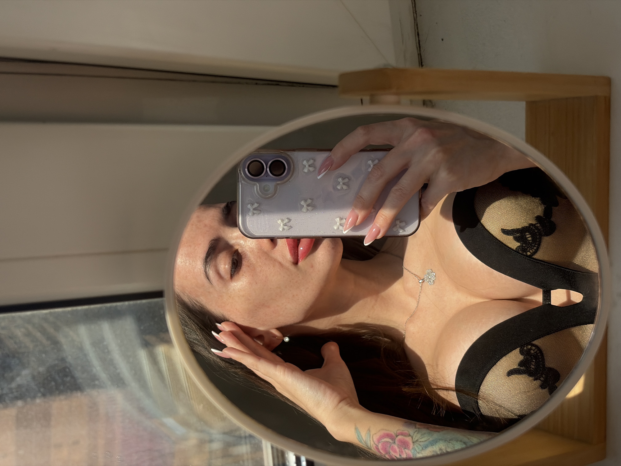 An image depicts a woman taking a mirror selfie indoors in Saint Petersburg, Russia. The woman is reflected from the chest up in a light-colored wooden-framed mirror. She has long dark hair, visible freckles, and is wearing red lipstick. Her left hand is raised to her temple, revealing a floral tattoo on her upper arm and a dangling clear stone earring on her right ear. A silver necklace with a clover pendant rests on her décolletage, above a black and beige sheer lace bra or bodysuit. Her right hand holds a light purple smartphone with a decorative bow-patterned case and dual camera lenses, angled towards the mirror to capture the reflection. Her nails are long, manicured with white French tips. Strong natural light illuminates her from the right, creating highlights. The reflection in the background shows a window with light filtering through, and a plain white wall is visible to the right of the mirror, with the mirror resting on a light-colored wooden surface.