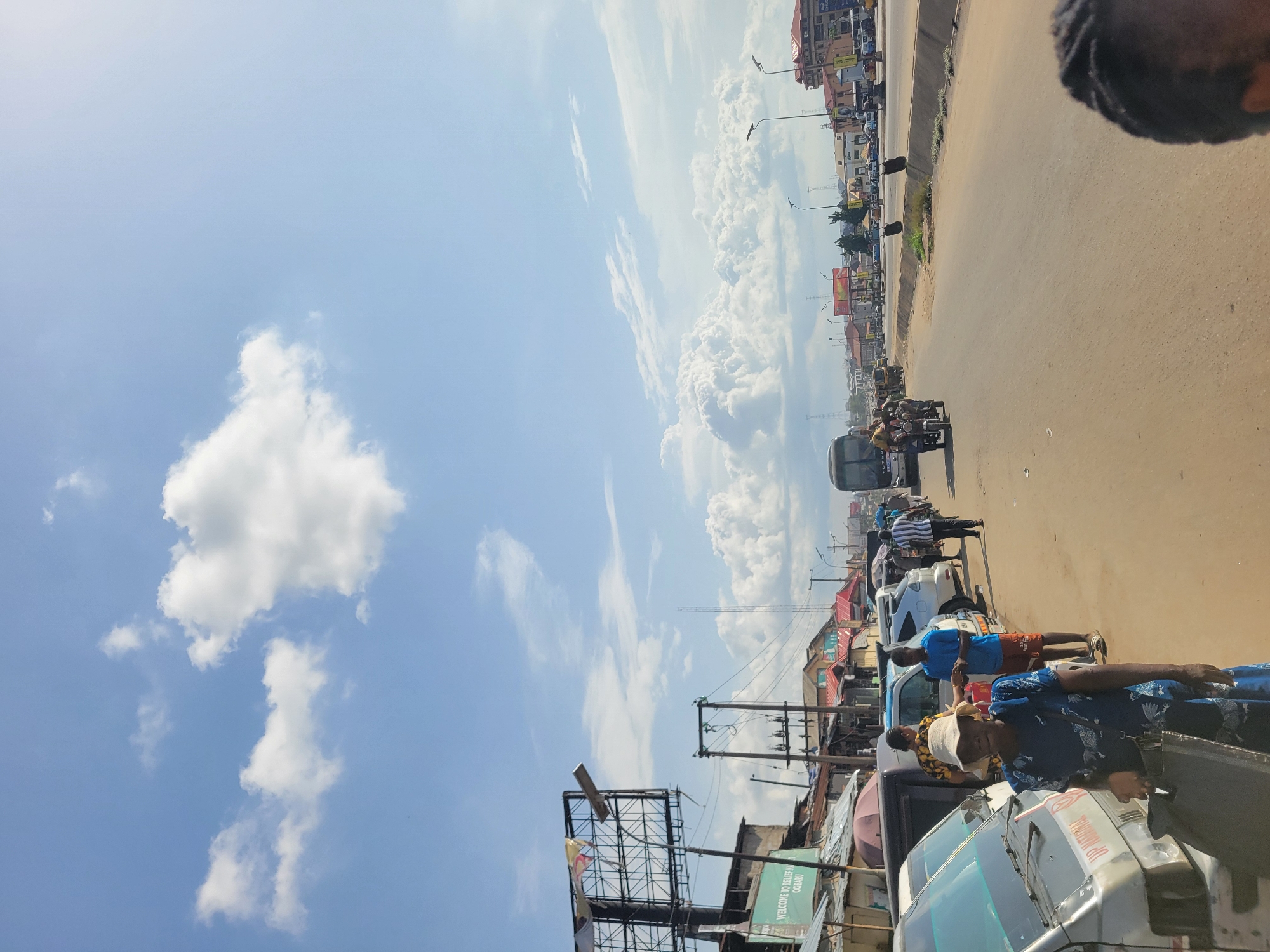 This media file captures a wide-angle street scene in what appears to be a bustling urban or semi-urban area, likely Fegge, Nigeria, during bright daylight hours. The weather is clear and sunny, featuring a vibrant blue sky adorned with large, fluffy white cumulus clouds that occupy a significant portion of the upper frame.
The foreground and midground are dominated by a wide, dusty or unpaved road that stretches into the distance. Along the right side of the road, a line of vehicles is visible, including various cars, a white bus-like vehicle, and motorcycles. Many people are present on and alongside the road. In the foreground, a woman wearing a blue patterned dress and a light-colored hat stands near a white van or bus, while another person in a blue shirt is near a white car further down. Closer to the background, a tricycle-like vehicle (often used for public transport) is seen on the road.
On the left side of the road, various structures line the street, including low-rise buildings with different roof styles, some featuring red corrugated iron. Power poles, electrical wires, and streetlights are also visible, suggesting developed infrastructure. At least two large, empty metal billboard frames stand prominently, one close to the foreground.
Visible text includes "WELCOME TO RELIEF M OGBAKU" on a green sign attached to a post near the road, and "UP NATIONAL" is printed on the side of the white bus-like vehicle in the foreground. The overall impression is one of active street life in a commercial or residential district under a clear sky.