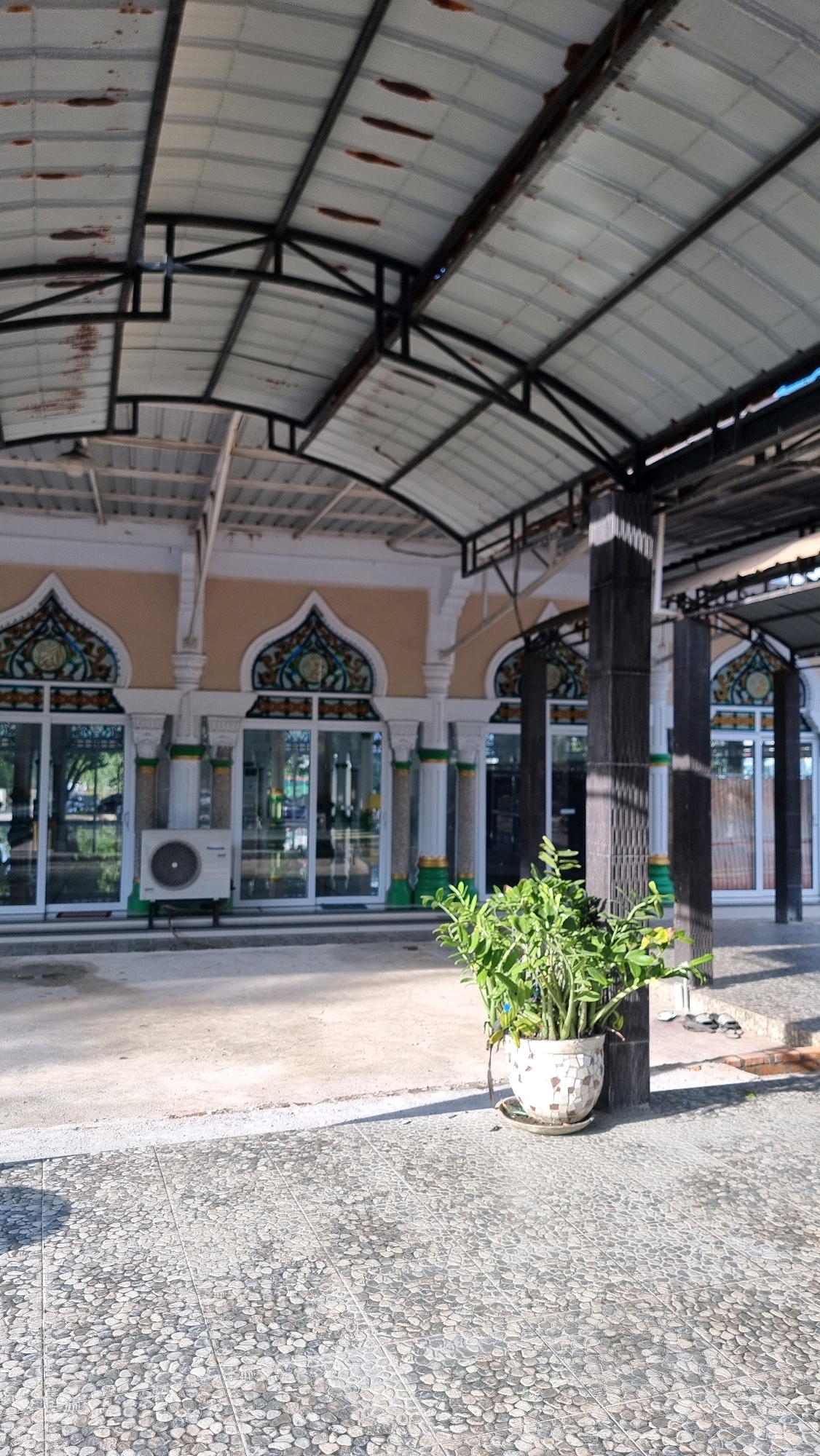 The image depicts the exterior of a building, possibly a mosque, in Blangrambong, Indonesia. The architecture features a curved roof supported by a metal frame, with some signs of rust on the roofing material. The facade of the building has large glass doors and windows, with decorative arched elements above some of them. There are white pillars with green accents. An outdoor air conditioning unit is visible to the left of the entrance. In the foreground, a potted plant with green foliage sits on a mosaic-patterned tiled surface. The ground in front of the building is a mix of concrete and the same tiled material. Sunlight casts shadows on the ground. The scene appears to be set in a public area, with no people clearly visible in the frame.