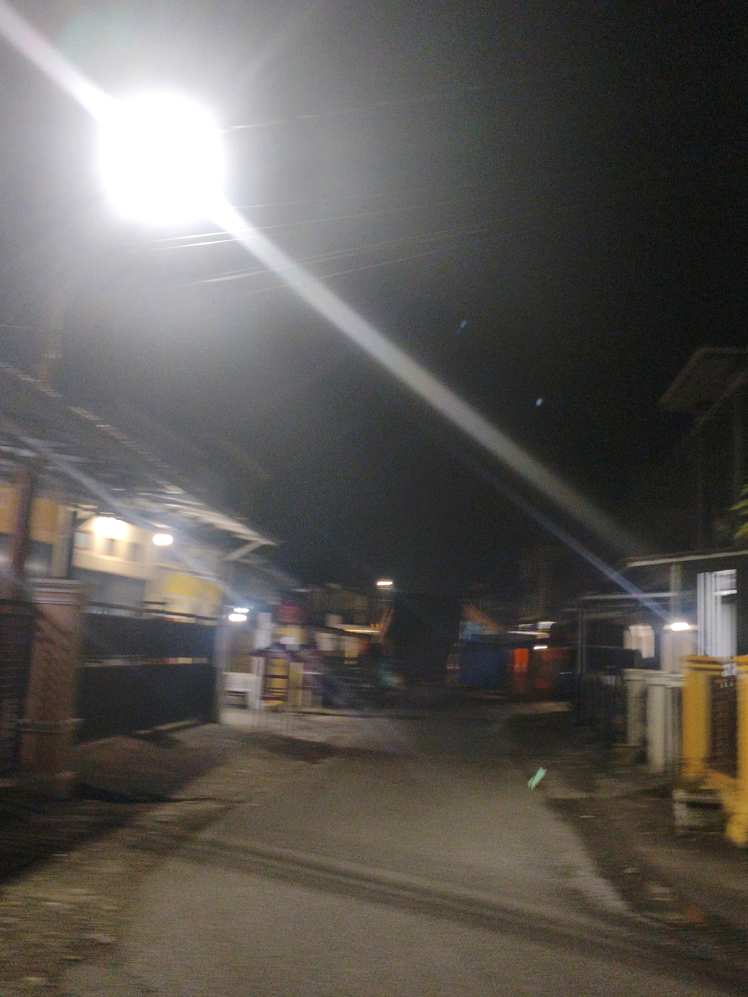 The image depicts a street scene at night in Koto Baru Semurup, Indonesia, illuminated by a bright overhead light source causing significant glare. The street appears to be paved, with a wet or reflective surface suggesting recent rain. On the left side of the street, there are structures with visible roofs and some illuminated windows, possibly shops or residences. Fences or walls are present along the property lines. On the right side, more buildings are visible, including one with a white vertical structure and some yellow elements, likely part of a building or fence. The overall impression is a somewhat blurry, dimly lit urban or semi-urban environment typical of a developing area at night. No people are clearly discernible in the image.