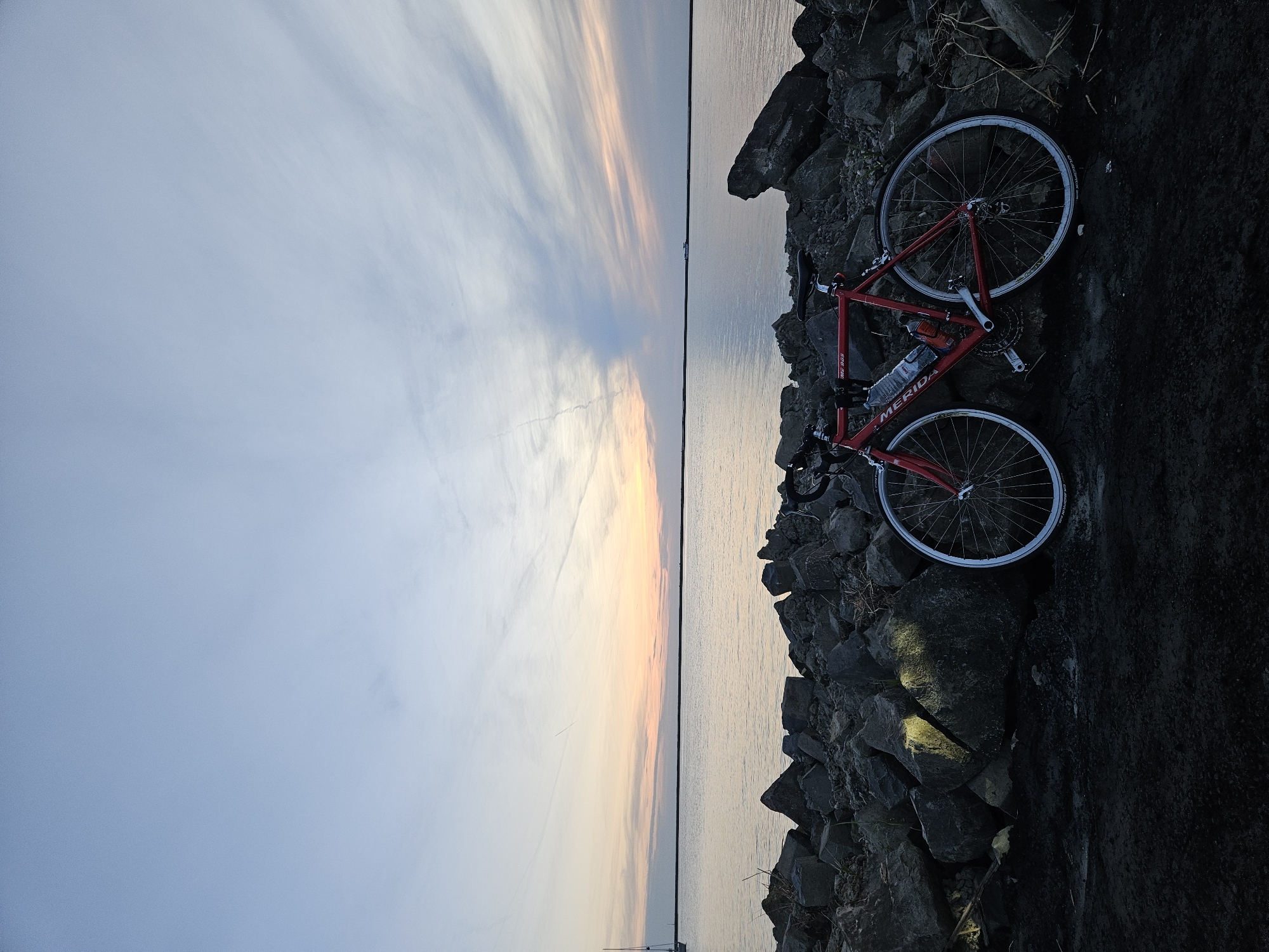 A red Merida road bicycle is leaning against a rocky embankment at the water's edge during sunset. The bicycle has two water bottles attached to its frame and white-rimmed wheels. The embankment is composed of large, dark grey rocks. The water stretches out to the horizon, reflecting the soft, pastel colors of the sky, which transitions from a pale blue at the top to hues of orange and pink near the horizon. Wispy clouds are scattered across the sky, with a visible contrail. In the distance on the left, a sailboat mast is visible. The scene suggests an outdoor recreational setting, possibly a dike or waterfront path, in or around Lelystad, Netherlands, given the characteristic landscape of the Flevopolder and its proximity to large bodies of water like the IJsselmeer. The overall atmosphere is serene and quiet, indicative of the end of the day.