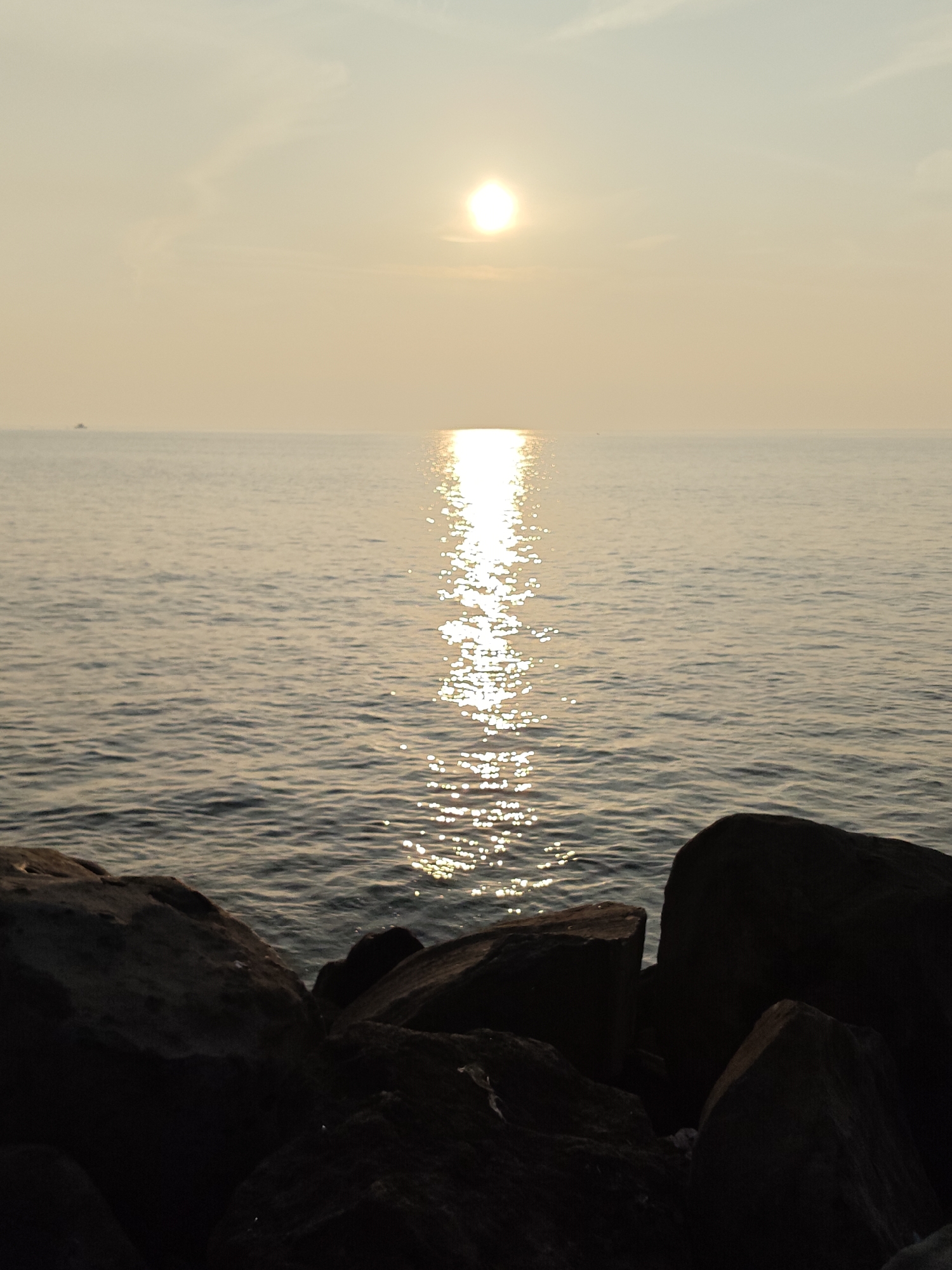 A serene sunset over the ocean is captured from a rocky shoreline. The sun, a bright orb, hangs low in the hazy sky, casting a shimmering golden path across the calm water. The sea reflects the soft hues of the sky, creating a tranquil atmosphere. In the distance, a small boat is visible on the horizon, adding a sense of scale to the vastness of the ocean. The foreground is dominated by dark, rugged rocks, providing a stark contrast to the luminous sky and water. The overall scene evokes a sense of peace and natural beauty. The image is believed to be taken in Lhokseumawe, Indonesia, a coastal city known for its beautiful beaches and sunsets.