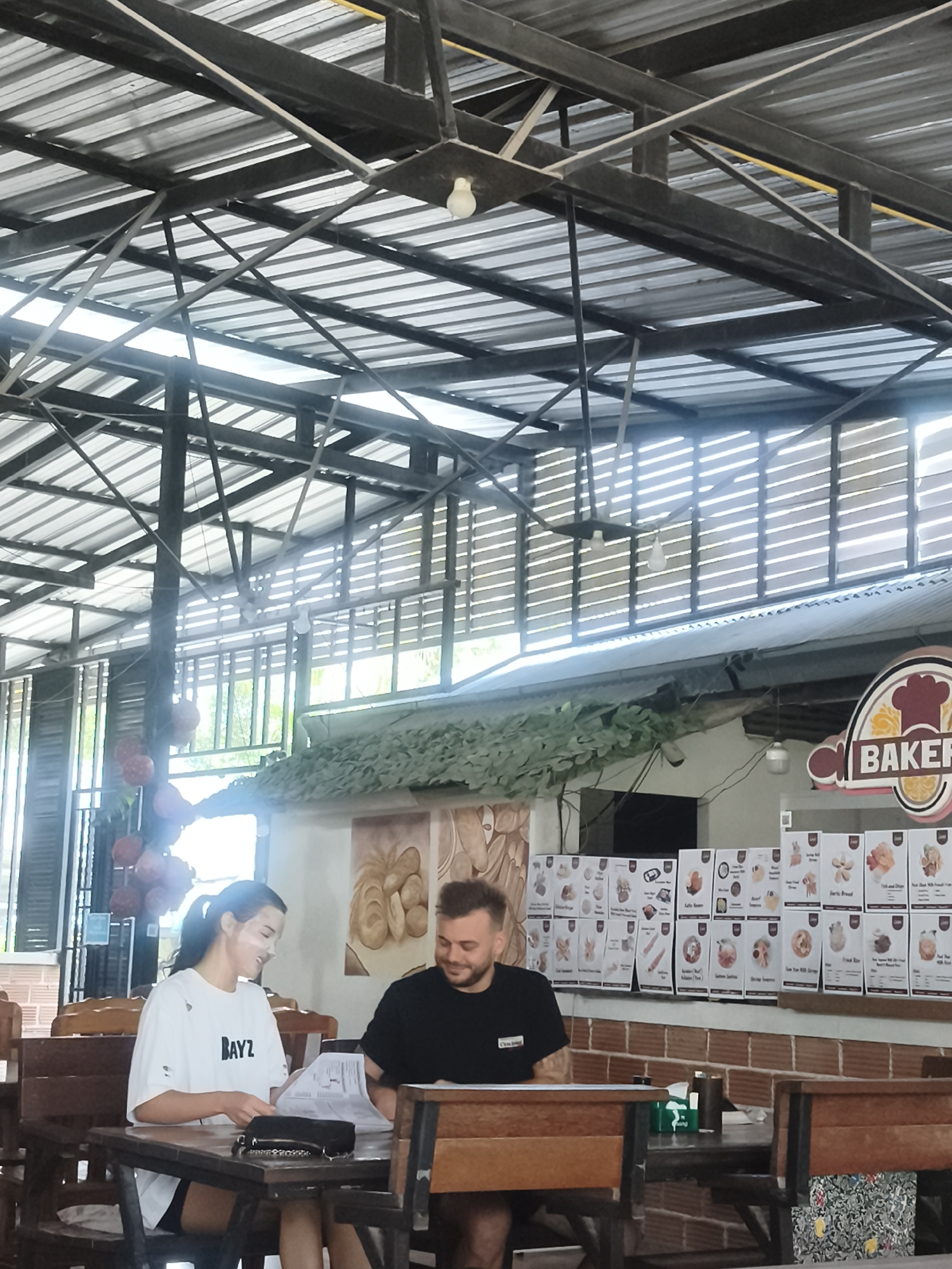 A photograph taken in Baan Mae Nam, Thailand, captures a man and a woman seated at a wooden table within an open-air structure. The woman, on the left, wears a white t-shirt with the word "BAYZ" printed on it, while the man, to her right, is dressed in a black t-shirt. They appear to be reviewing documents placed on the table between them.
Behind them, a bakery is visible, indicated by the "BAKER" sign. Menu items with pictures are displayed along the brick wall. The structure features a metal roof with an elaborate framework of beams and supports. Light fixtures hang from the ceiling structure. The environment suggests a casual dining setting.