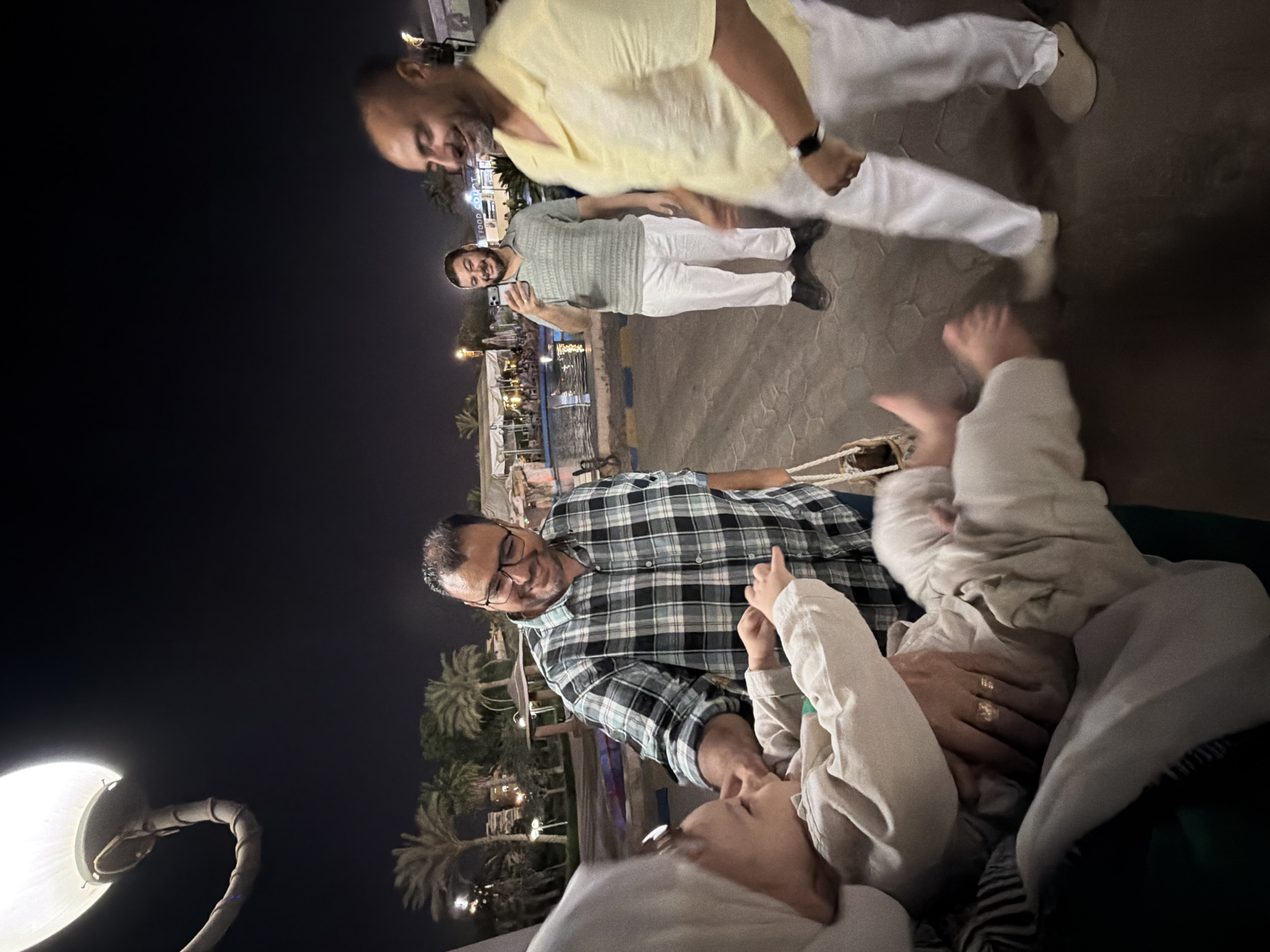 The image depicts a nighttime scene in a public outdoor area, possibly a restaurant or entertainment venue, in the city of 6th of October, Egypt. In the foreground, a man in a plaid shirt is interacting with a baby who is being held. The baby is wearing a light-colored outfit and appears to be reaching out. The man has glasses and is smiling. To the right of the man in the plaid shirt, another man in a yellow short-sleeved shirt and white pants is walking past, looking to his right. In the background, a third man is standing, holding a mobile phone and appearing to take a photograph or video. He is wearing a green patterned shirt and white pants. Behind the people, there is a body of water, possibly a swimming pool, with seating areas and lights in the distance, suggesting a leisure or dining establishment. Palm trees and some buildings are visible in the dimly lit environment. The ground is paved with patterned stones. A large lamp post with a bright light is visible in the upper left corner of the frame.