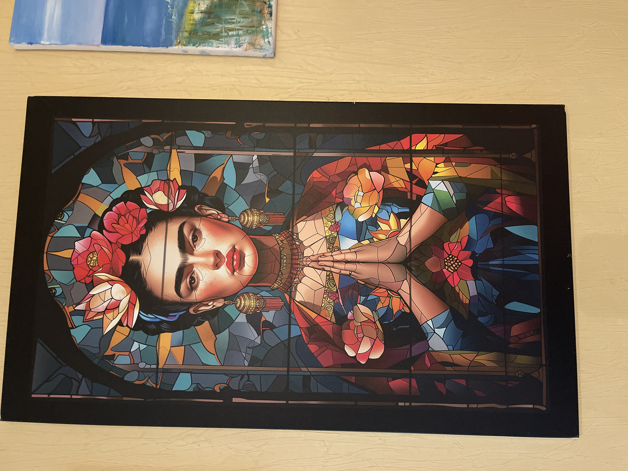 The image shows a stylized portrait of Frida Kahlo, depicted in a manner reminiscent of stained glass artwork. She is positioned horizontally, with her hands clasped in front of her chest. Her signature floral headdress is prominently displayed, and she wears traditional Mexican jewelry. The background consists of a mosaic of colorful, geometric shapes that suggest stained glass panes. The artwork is framed in a black border. It is positioned on a table. Another painting is partially visible at the top.