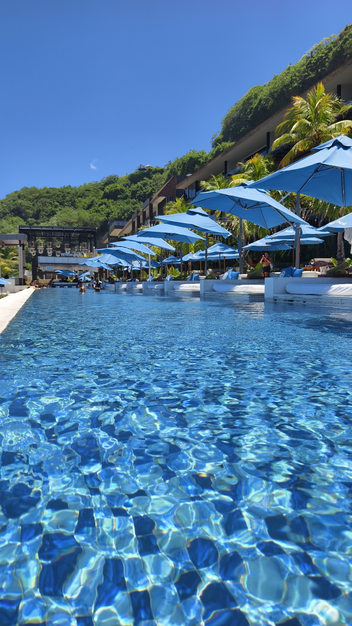 The image shows a swimming pool at a resort in Kuta Selatan, Indonesia. The pool water is clear and blue, with patterns of light reflecting off the bottom. Along the pool's edge are a series of white lounge beds with blue umbrellas. People are visible in and around the pool, including someone standing near one of the lounge areas. The resort is built into a hillside covered in green vegetation. A building with multiple levels and what appears to be a bar or restaurant area is also visible behind the pool and lounge area. The sky is clear and blue.