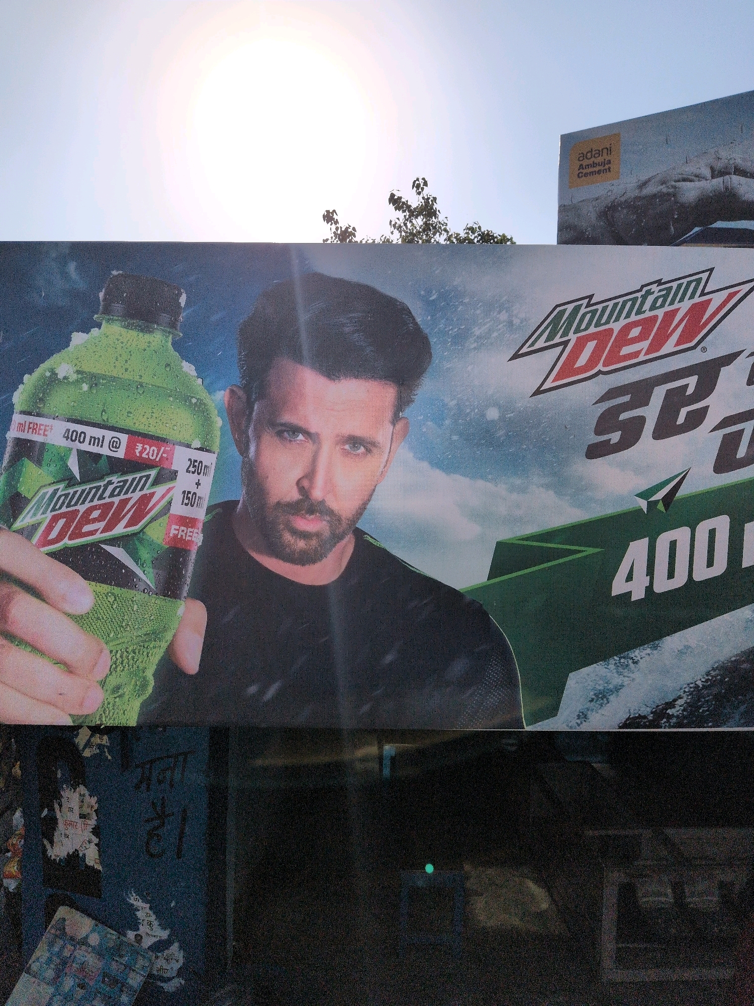 The image shows a billboard advertising Mountain Dew, featuring actor Hrithik Roshan holding a bottle of the beverage. The billboard is large and prominently displayed, suggesting it's a public advertisement. In the background, another billboard for "adani Ambuja Cement" is visible, indicating a commercial area. The sky is bright and sunny, with the sun's rays casting a strong glare. The lower portion of the image shows a dark, possibly street-level view with some posters and graffiti on a blue surface. The presence of posters and graffiti suggests an urban or semi-urban setting. There is a small green light visible in the dark lower section, possibly from a street light or a business. The overall scene appears to be an outdoor advertisement in a bustling area. Given the mention of "adani Ambuja Cement," which is an Indian company, and the script on the posters in the lower section appearing to be Hindi, it is likely located in India. While the exact location in Chetru, India, cannot be definitively determined from the image alone, it is consistent with an Indian urban or commercial landscape.