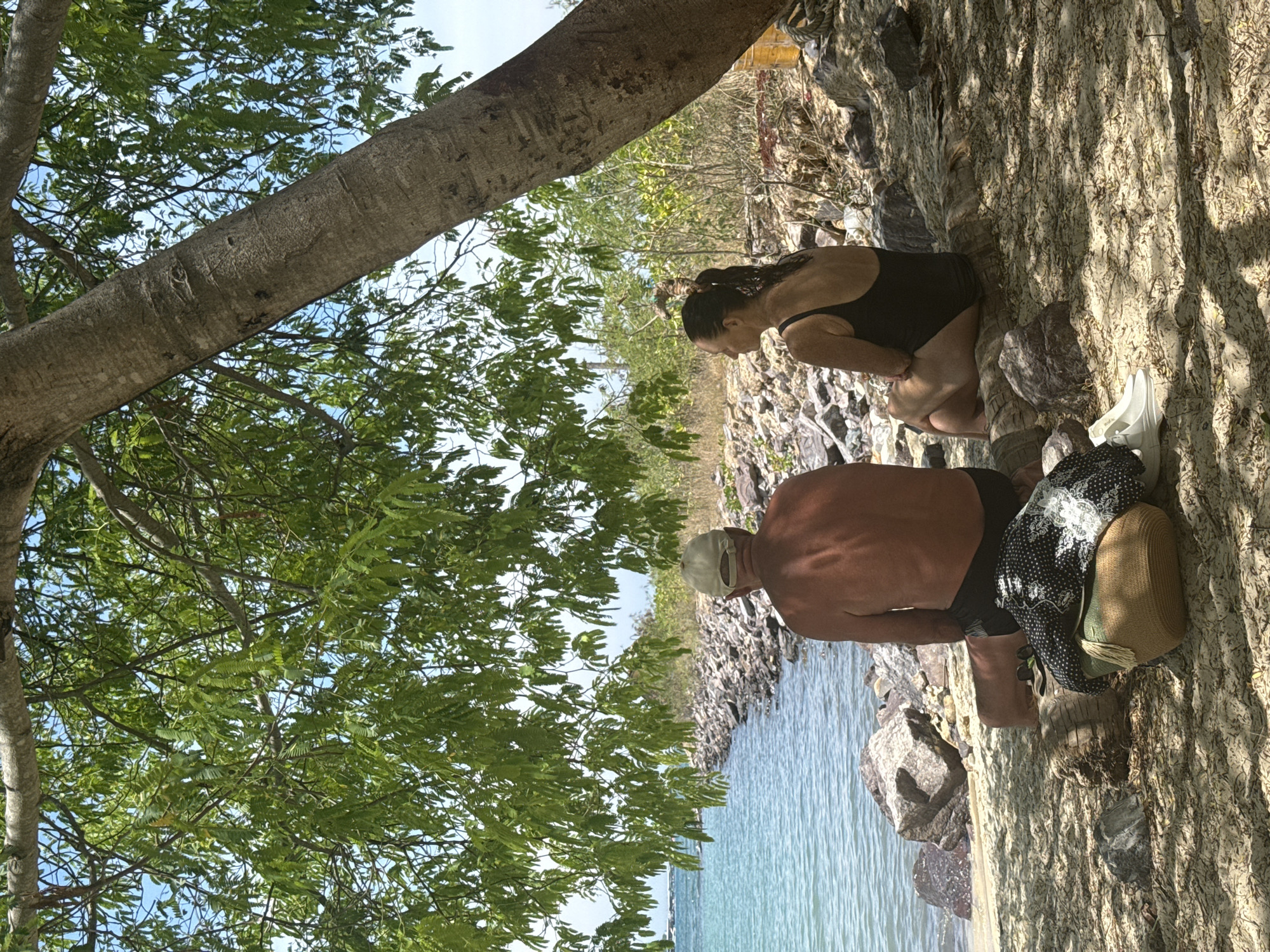 The image depicts a man and a woman sitting on a beach, shaded by a large tree. The man, wearing a cap and dark shorts, faces away from the camera towards the turquoise water. The woman, in a black swimsuit, sits beside him, also facing the water. In the foreground, a straw bag with a patterned garment draped over it and a pair of white sneakers are visible on the sandy ground. The background shows a rocky shoreline and sparse vegetation under a bright, clear sky. The scene is set at a beach location, potentially in or near the city of Nha Trang, Vietnam, given the presence of tropical foliage and the coastal environment.