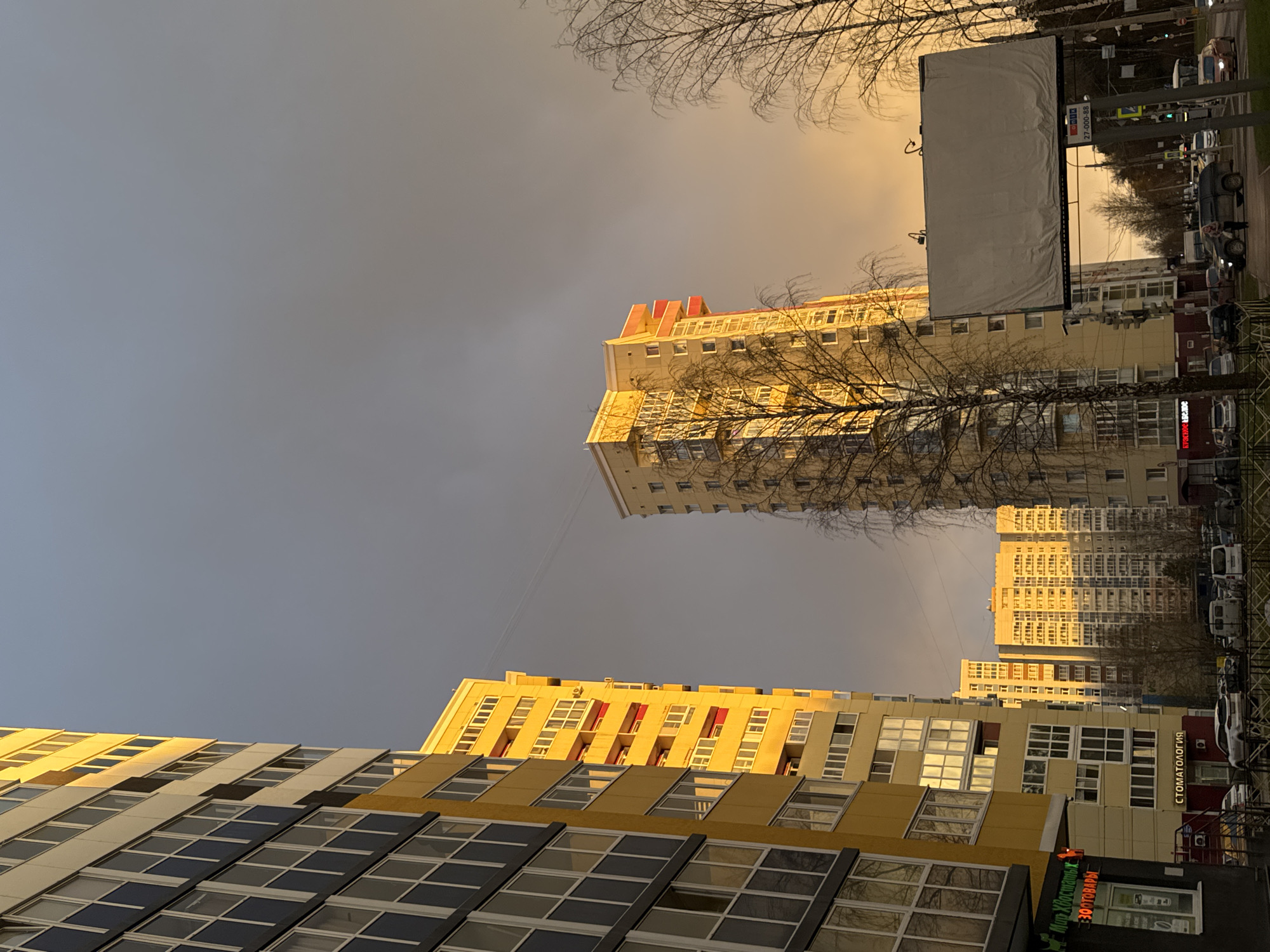 The image captures an urban residential scene in Perm, Russia, during the golden hour of either sunrise or sunset. The sky is dramatically split, with a dark grey, almost stormy-looking area on the left gradually transitioning to a bright, golden-orange hue on the right where the sun's low rays are breaking through.
Several modern, multi-story apartment buildings dominate the landscape. In the mid-ground, a tall, light-colored building with red accents on its upper floors is bathed in the warm, golden sunlight, causing its many windows to glow. Bare tree branches, typical of winter or early spring, are silhouetted against the bright sky and buildings, adding a stark, natural element.
In the foreground, another contemporary building features a facade with alternating sections of yellow, grey, and dark window panels, also brightly illuminated by the golden light. This building includes retail spaces on its lower levels.
To the right, a street runs parallel to the buildings, with several cars parked along the curb and a few in motion, suggesting ongoing daily life. A large, blank billboard stands prominently beside the street, featuring a metal frame. Power lines or cables are faintly visible stretching across the sky.
Visible text on the buildings includes a red sign that reads "СТОМАТОЛОГИЯ" (Dentistry) and, below it, a green sign with "Мир Хлопотычи" and "ЗООТОВАРЫ" (Pet Supplies). The billboard displays a phone number "27-000-888" and "МЕНЮ" (Menu) faintly.
The scene conveys a typical city environment under striking natural lighting, highlighting the modern architectural style and the contrast between the illuminated buildings and the varied sky.