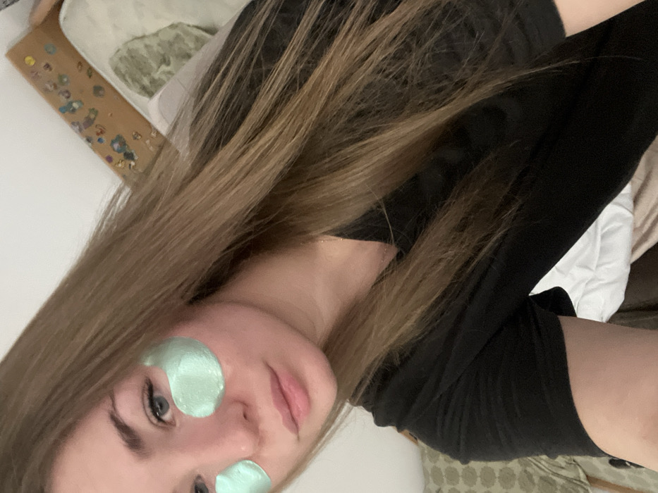The image captures a close-up portrait of a young person, likely female, with long, straight, light brown hair that cascades over their shoulders and chest. The photo is taken at a tilted angle, with the person looking directly into the camera. They have light-colored eyes (appearing blue or grey), light eyebrows, and a fair complexion. Their lips have a natural pink tint, and they exhibit a relaxed, neutral expression.
The most prominent detail on their face is the presence of two mint-green, gel-like eye patches, one applied directly under each eye, indicating a skincare or self-care routine. The person is wearing a black, long-sleeved top or shirt.
The background suggests an indoor setting, possibly a bedroom. To the upper left of the frame (relative to the person's head), there's a light-colored surface and a brown object adorned with small, colorful, possibly glittery or sequined embellishments. To the right side of the frame, behind the person's shoulder, a white fabric, likely a pillow or duvet, is visible, along with a glimpse of a patterned fabric (in shades of green and brown) further in the background. The lighting is soft and ambient. No text is visible in the image.