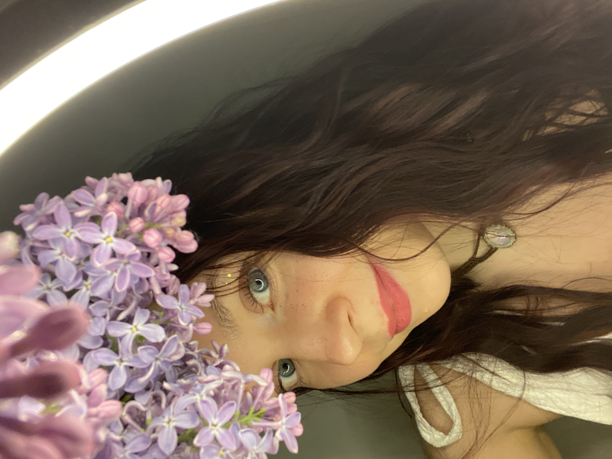 A selfie-style image features a person with pale skin, light blue eyes, and freckles visible on their nose. Their dark brown, wavy hair fills a significant portion of the frame, especially towards the top right. The person is positioned at an angle, with their face angled towards the viewer. They are wearing a white top with thin straps. A cluster of purple lilac flowers is prominently displayed in the lower-left corner of the image. A soft, circular light source, possibly a ring light, is visible in the upper-left corner. The person has a smear of red lipstick on their cheek. A necklace with a light-colored pendant is visible on their neck. The location is unknown, not determined to be Chaikovsky, Russia.