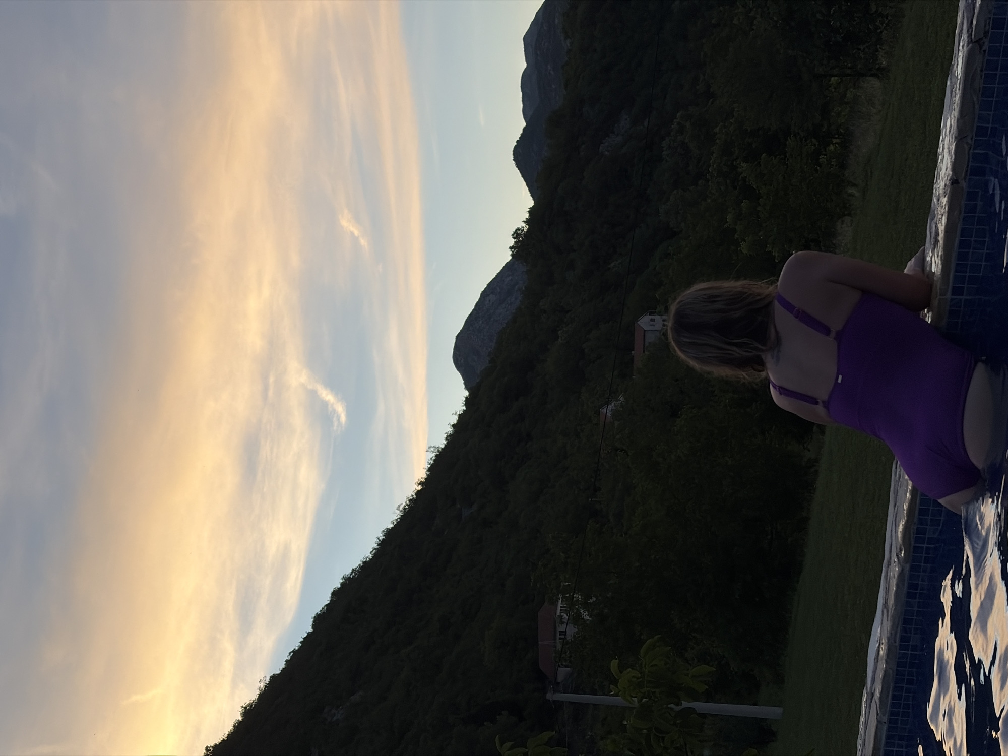An over-the-shoulder shot captures a woman in a purple one-piece swimsuit resting her arms on the edge of an infinity pool. She is positioned in the lower right corner of the frame, facing away from the camera and towards a scenic mountainous landscape. The pool water reflects the soft hues of the sky. The background is dominated by a steep, tree-covered mountainside, with a few houses nestled within the foliage. The sky above transitions from a pale yellow and orange near the horizon, suggesting either sunrise or sunset, to a soft blue higher up, with wispy clouds scattered across it. The overall impression is one of serene contemplation in a natural, possibly rural, setting, likely in or around Ceklin, Montenegro.