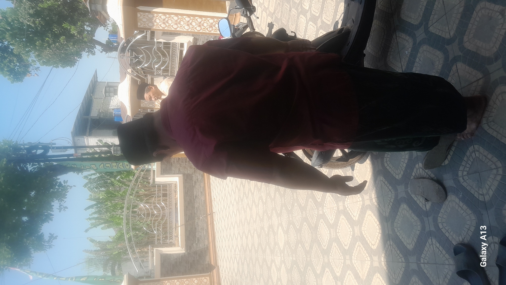 A medium shot captures an adult male from behind, walking away from the camera on a tiled outdoor patio. He wears a dark maroon collared shirt and dark green pants. His head is covered by a black cap. To his right, a portion of a dark-colored motorcycle is visible. In the background, a man in a light-colored shirt and black cap is walking towards the camera. Behind him, a building with a white facade and tiled roof stands under a clear blue sky. A fence with ornate metalwork separates the patio from the building. The ground is covered in diamond-patterned light-colored tiles. Several pairs of sandals are scattered on the ground. The scene appears to be in Hadipolo, Indonesia, based on the "Galaxy A13" watermark which suggests the image was taken with a Samsung phone, commonly used in the region.