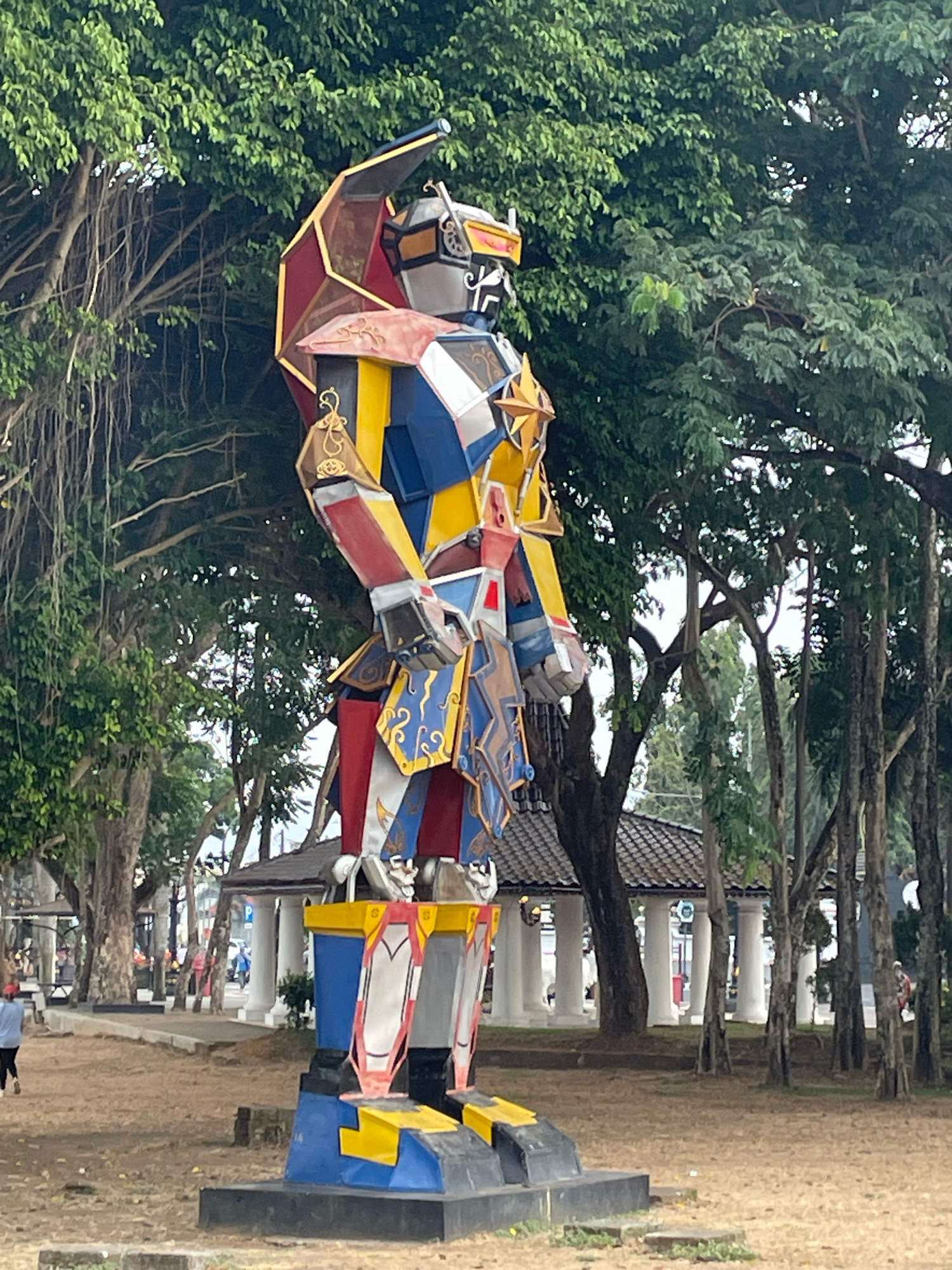 In Purworejo, Indonesia, a large, colorful statue of a robot stands prominently in an outdoor park. The robot is intricately designed with a striking combination of blue, yellow, red, and white panels, featuring ornate gold accents and a prominent red and gold helmet-like structure on its head. It appears to be a stationary, decorative art piece. The background reveals lush green trees with hanging vines, and a paved walkway lined with trees and buildings. In the distance, there is a white gazebo-like structure with white columns and a tiled roof. Several people are visible in the background, walking and engaged in outdoor activities, suggesting a public recreational area. The ground is covered with dry soil and scattered leaves.