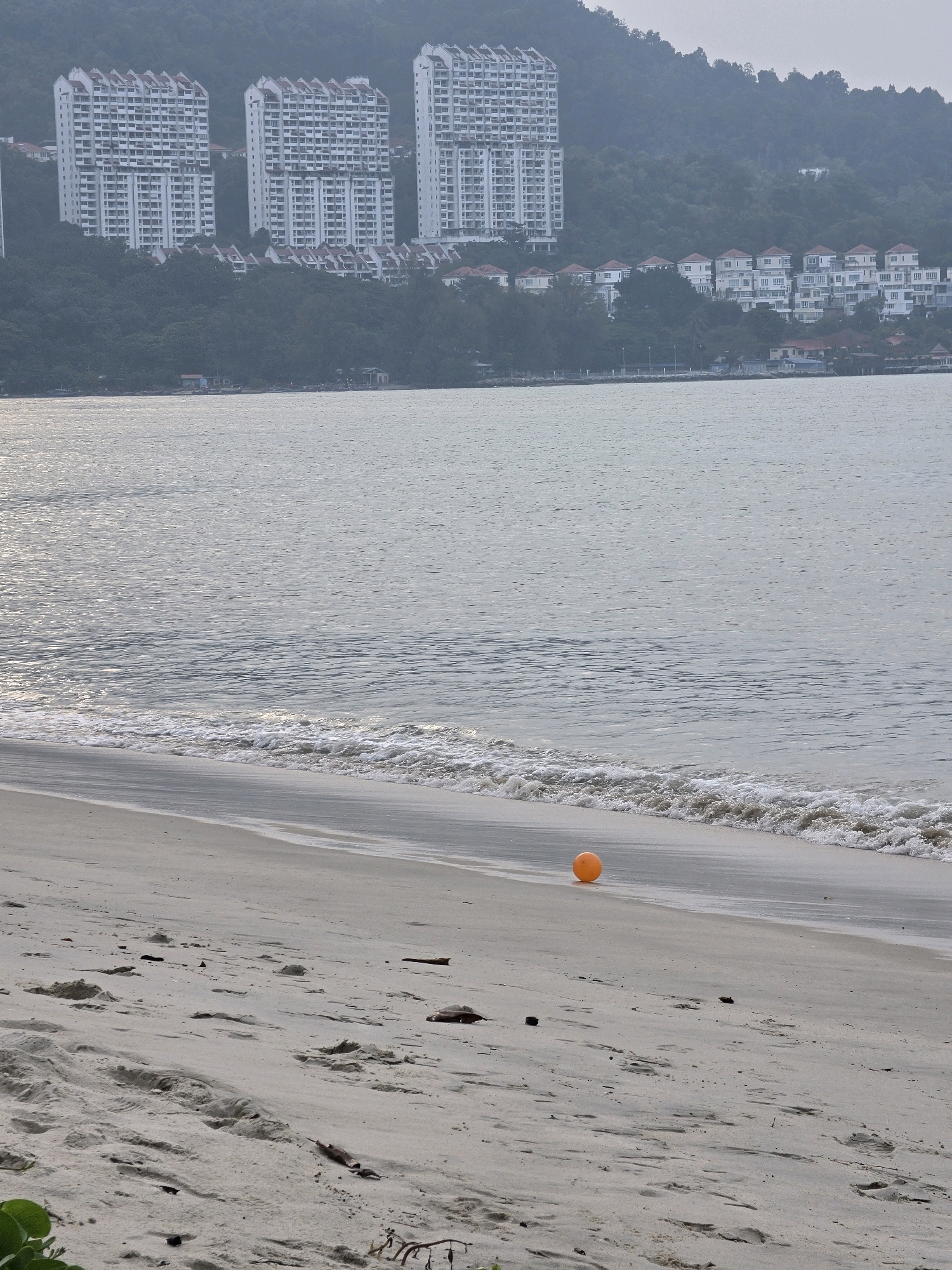 The image depicts a beach scene with the city of George Town, Malaysia, visible in the background. Several high-rise buildings, characteristic of modern architecture, are situated on a hillside overlooking the water. The beach itself is sandy, with gentle waves lapping at the shore. An orange ball is resting on the sand near the water's edge. The overall atmosphere is serene, with the overcast sky casting a soft light on the landscape. The scene suggests a leisurely day at the beach, with the natural beauty of the coastline complementing the urban development in the distance.