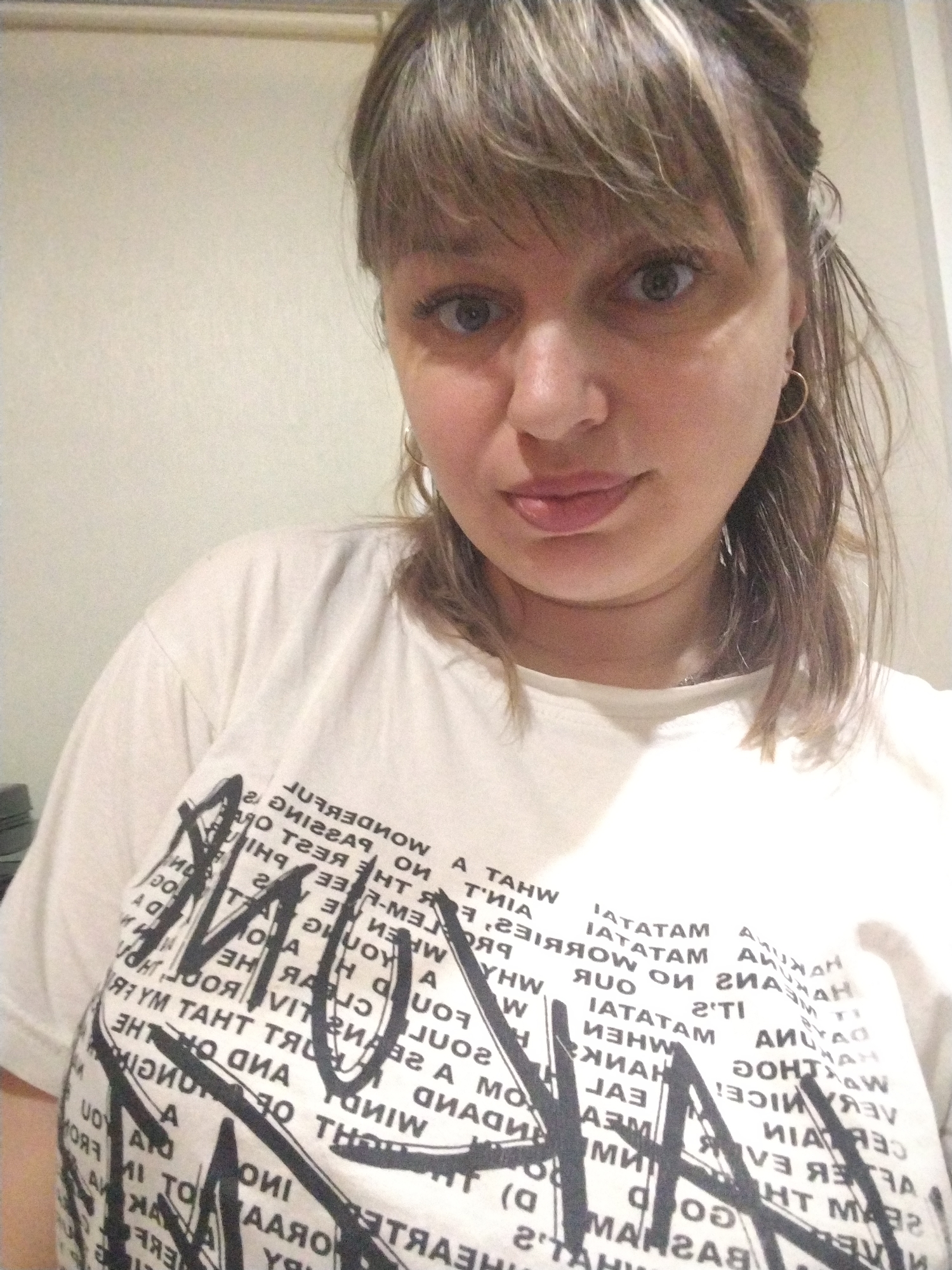 A medium shot shows a Caucasian woman with light brown hair and bangs. She is wearing gold hoop earrings and a white T-shirt with black, stylized writing. The writing appears to be an artistic design incorporating words and phrases. The background is a plain, light-colored wall. There is no contextual information in the image to confirm or deny the location.