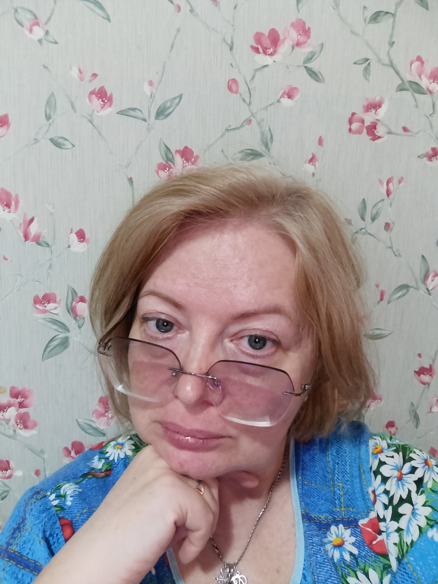 A woman with short, blonde hair and blue eyes is posing for a photo. She is wearing glasses and a blue floral shirt with white daisies and red accents. Her left hand is supporting her chin, and she is wearing a silver necklace with a pendant. The background is a wallpaper with a pattern of pink flowers and green leaves. The photo appears to have been taken in a residential setting, potentially in the city of Большой Букор, Russia.