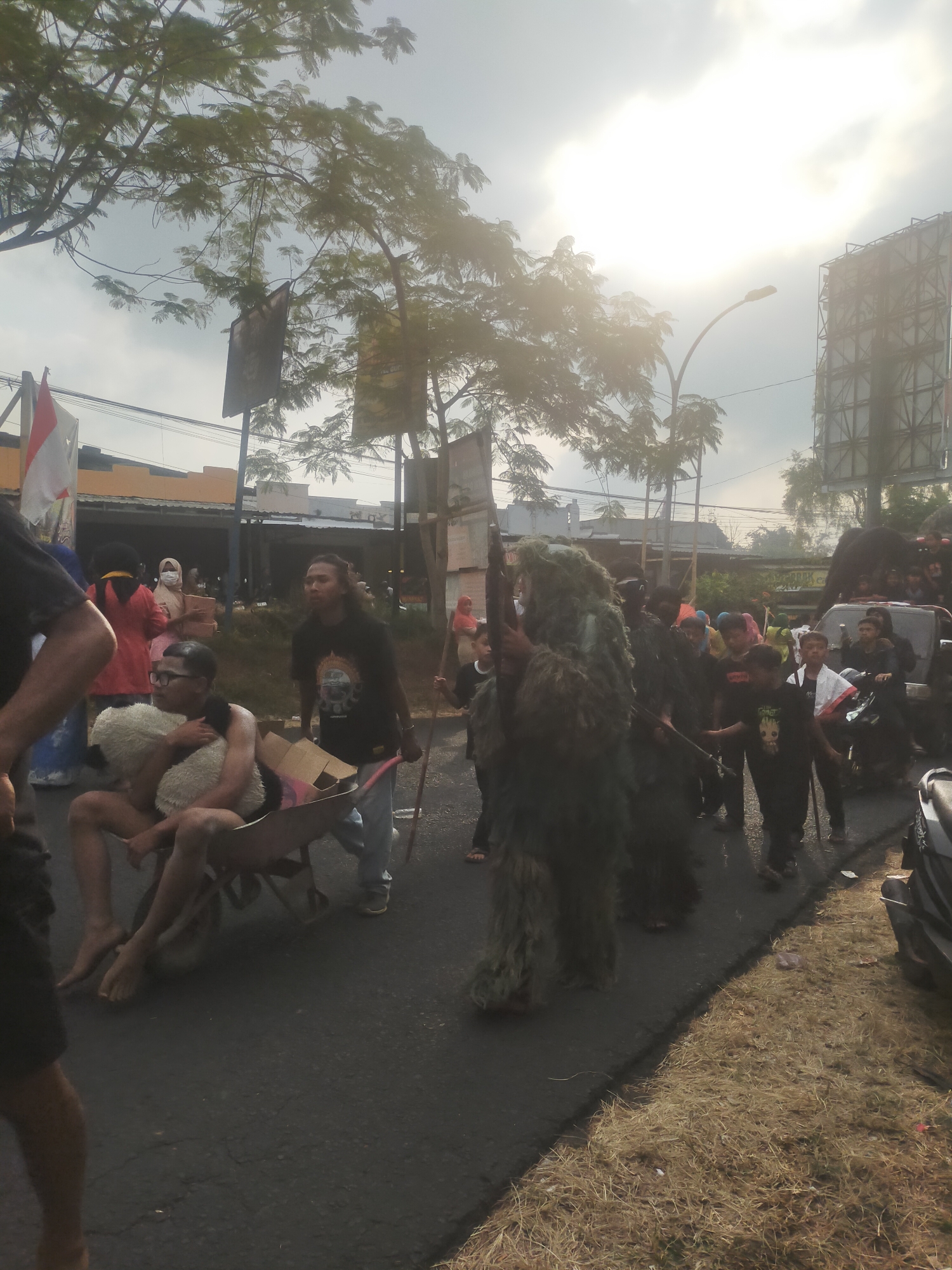 This is a medium-shot, eye-level, outdoor photograph taken on a street in Tuwel, Indonesia. The scene depicts a parade or procession. In the foreground, a person wearing a camouflage ghillie suit, holding a rifle, walks on the right side of the road. To the left, a person in a wheelbarrow is being pushed by another individual; the person in the wheelbarrow appears to be holding a fluffy object. Several other people are walking in the procession, some of whom are children. In the background, there are buildings, trees, and a billboard. The sky is overcast with bright sunlight filtering through. The road is paved and wet in some areas.