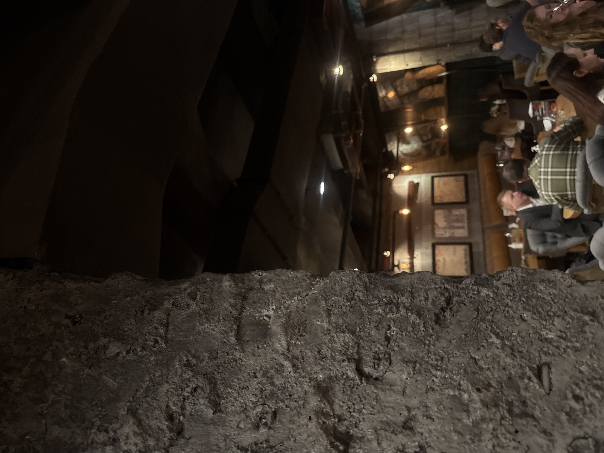 The image presents a dimly lit, rustic interior scene, potentially in a restaurant or pub in Yekaterinburg, Russia, captured from an elevated perspective looking downwards. The immediate foreground, occupying the lower half of the frame, is dominated by a rough, textured, grey-brown surface, resembling a stone wall or concrete barrier.
Beyond this foreground element, a vibrant, if subdued, social setting is visible. The establishment is characterized by warm, artificial lighting from several exposed bulb fixtures, illuminating dark wooden structures, possibly beams or paneling, and walls adorned with framed pictures.
Multiple people are present, seated at tables, appearing to be dining or socializing. Notable figures include an individual wearing a green and white plaid shirt, seen from behind, and another in a dark suit jacket, also facing away from the camera. Other patrons are visible further in the background, including a person with curly hair at the upper right. Tables are set with drinks and possibly meals.
The overall ambiance is cozy and intimate, suggesting an evening or nighttime setting. No visible text is discernible on any objects or surfaces within the frame. The unusual vantage point, looking over a substantial barrier, implies the shot was taken from a balcony, mezzanine, or a higher section of the venue.