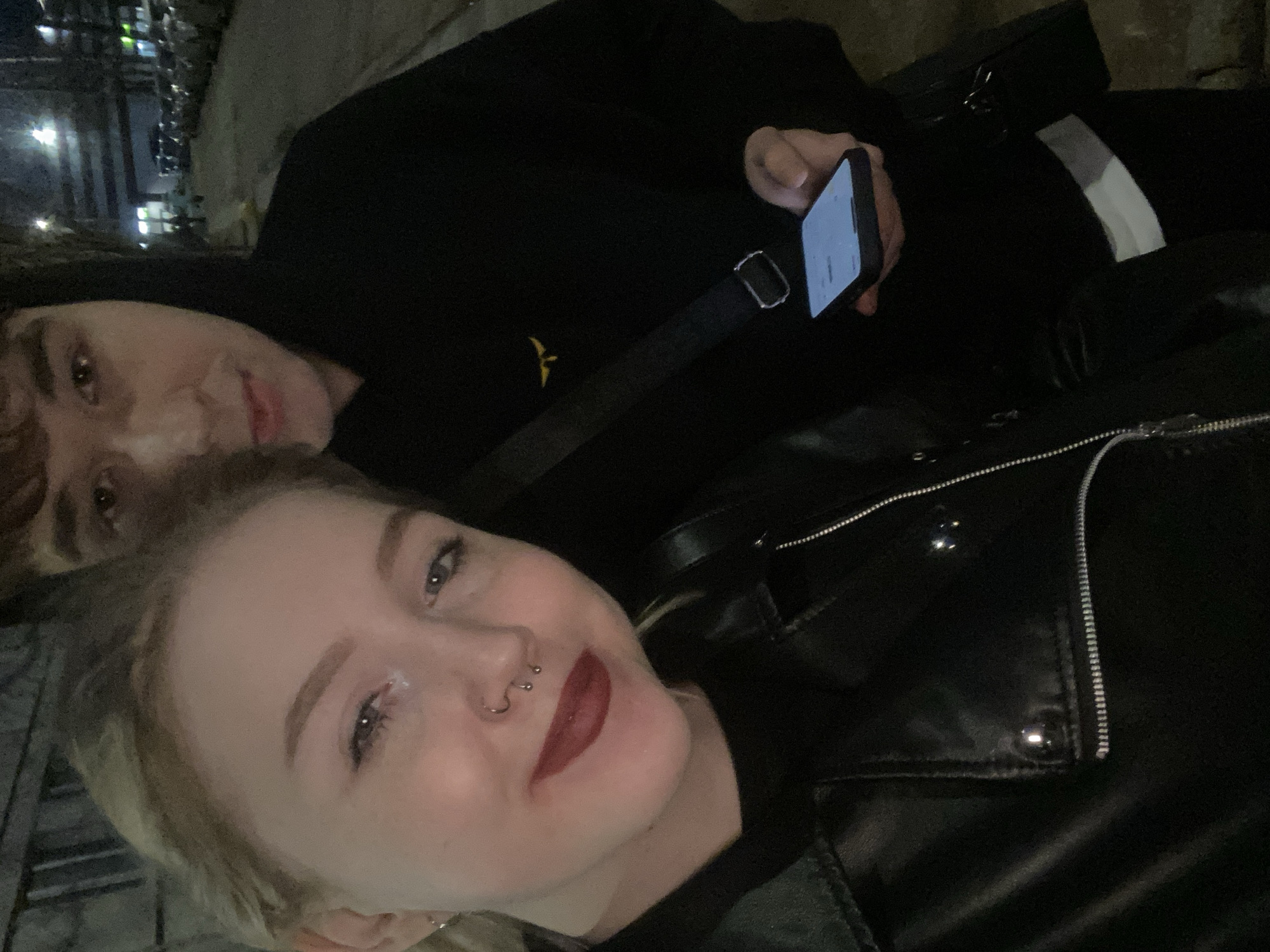 Two people are posing for a selfie outdoors at night, with streetlights visible in the background. The person in the foreground is a light-skinned woman with blonde hair, wearing dark red lipstick, and sporting piercings on her nose. She is wearing a black leather jacket. Behind her is a light-skinned man with brown hair, looking at the camera. He is wearing a black hoodie. He is holding a smartphone in his hand. A black bag is strapped across his body. The location is unknown, but specified as the city of Izhevsk, Russia.