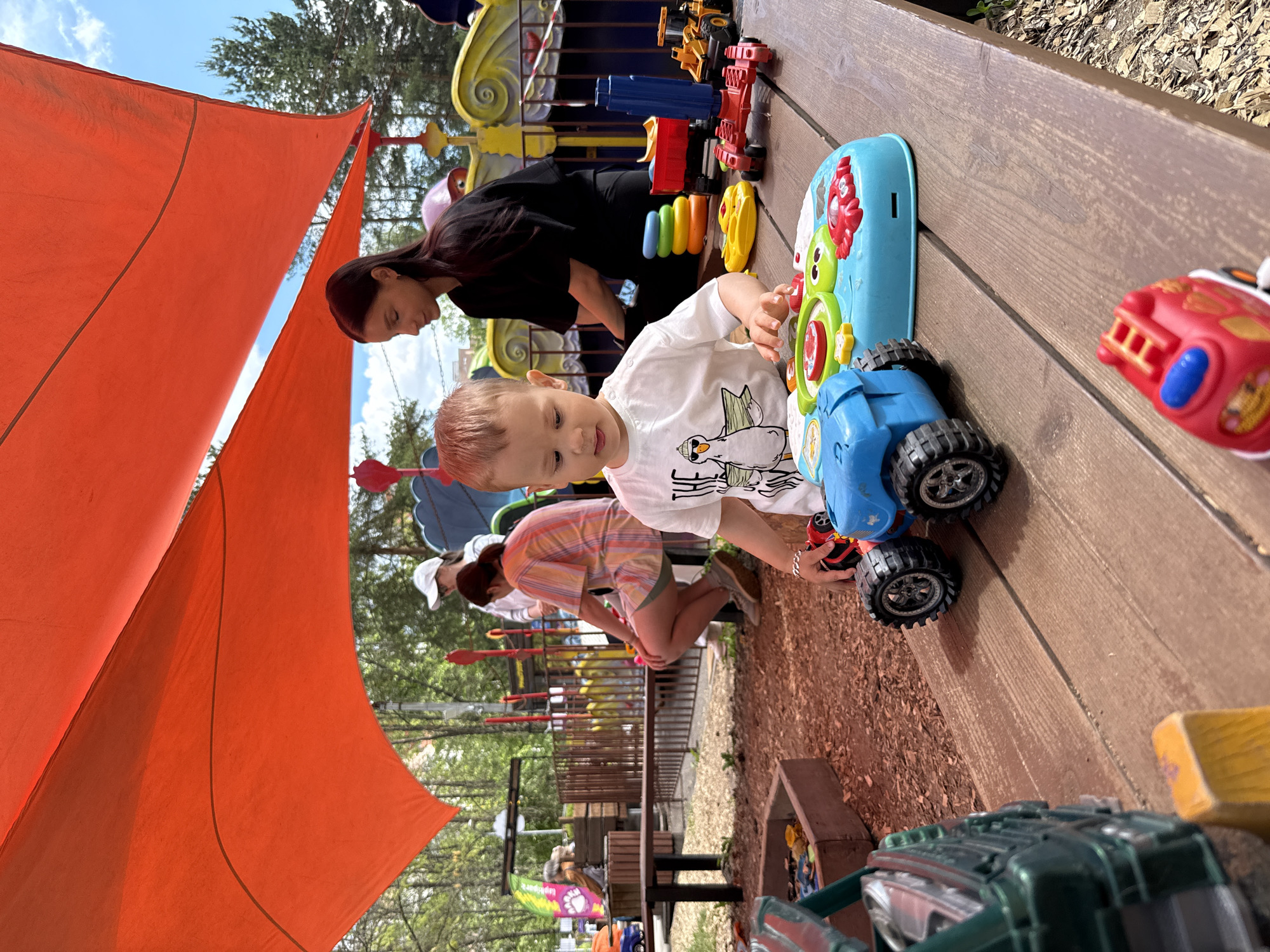 This outdoor scene captures a vibrant play area on a bright, sunny day, with the image rotated counter-clockwise. In the foreground, a young child with light brown hair, wearing a white t-shirt featuring a cartoon duck, is engrossed in playing with colorful toys on a wooden surface. He is focused on a blue and green electronic toy and a blue toy truck. Behind him, a woman with long dark hair, dressed in a black top, leans over the same wooden platform, observing the child. Further back, another person in a striped top and white cap is partially visible, kneeling amidst more toys.
The setting appears to be a well-equipped play space, possibly a park or daycare. A large, bright orange shade sail dominates the left side of the frame, providing cover. The ground below the play area is covered in wood chips. The wooden platform holds an assortment of toys, including construction vehicles like an excavator and a dump truck, a red toy fire truck, and a multi-colored stacking ring toy. Lush green trees are visible in the background against a clear blue sky with some white clouds, indicating pleasant weather. The child appears content and engaged in play. There is no visible text in the image.
Location context: Unknown, Unknown