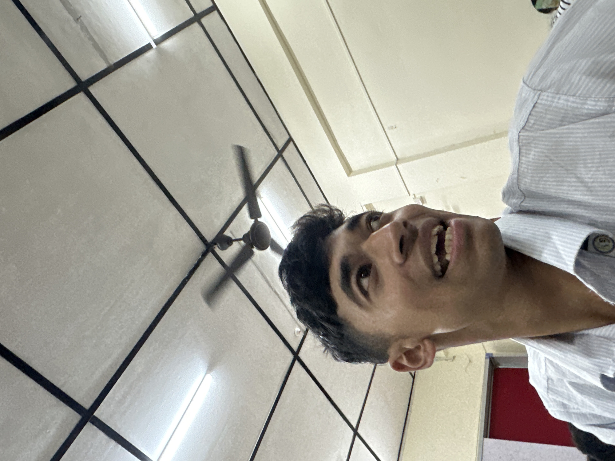 A young male with dark hair and brown eyes is looking upwards and to the right of the frame with a wide open mouth, revealing his teeth. He is wearing a light blue and white striped collared shirt. The background consists of a white ceiling with a black grid pattern and fluorescent lighting. A ceiling fan is visible in the upper portion of the image, with its blades blurred due to motion. To the left of the male, a portion of a red door is visible. The overall environment appears to be indoors, possibly an office or a classroom.