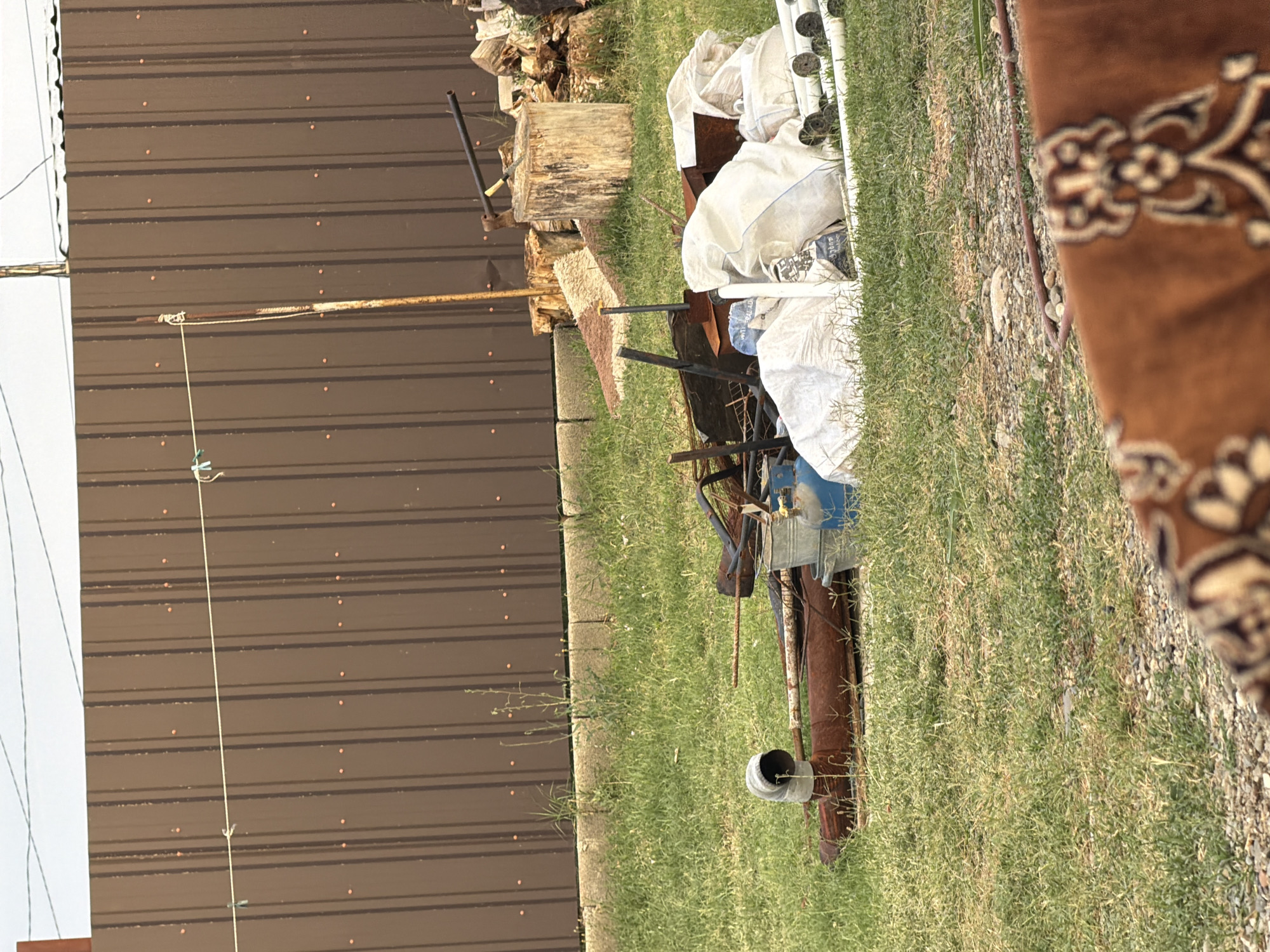 In the city of Tbilisi, Georgia, a collection of discarded items is situated on a grassy area next to a brown corrugated metal fence. The fence, with vertical seams and evenly spaced orange dots, stretches across the background. A thin rope, supported by a rusty metal pole and held by two clothespins, runs horizontally in front of the fence.
In the foreground, a pile of miscellaneous objects is scattered on the grass. These include rusted metal pipes, a gray bucket, a blue gas cylinder, and several large white plastic bags filled with unknown contents. To the right, a chopped wooden log sits with an axe embedded in it, and a stack of firewood is visible behind it. A small portion of a brown rug with an ornate pattern is also in the lower right corner of the image. The environment is outdoors, with green grass covering the ground and a hazy sky above. No people are visible in the image.