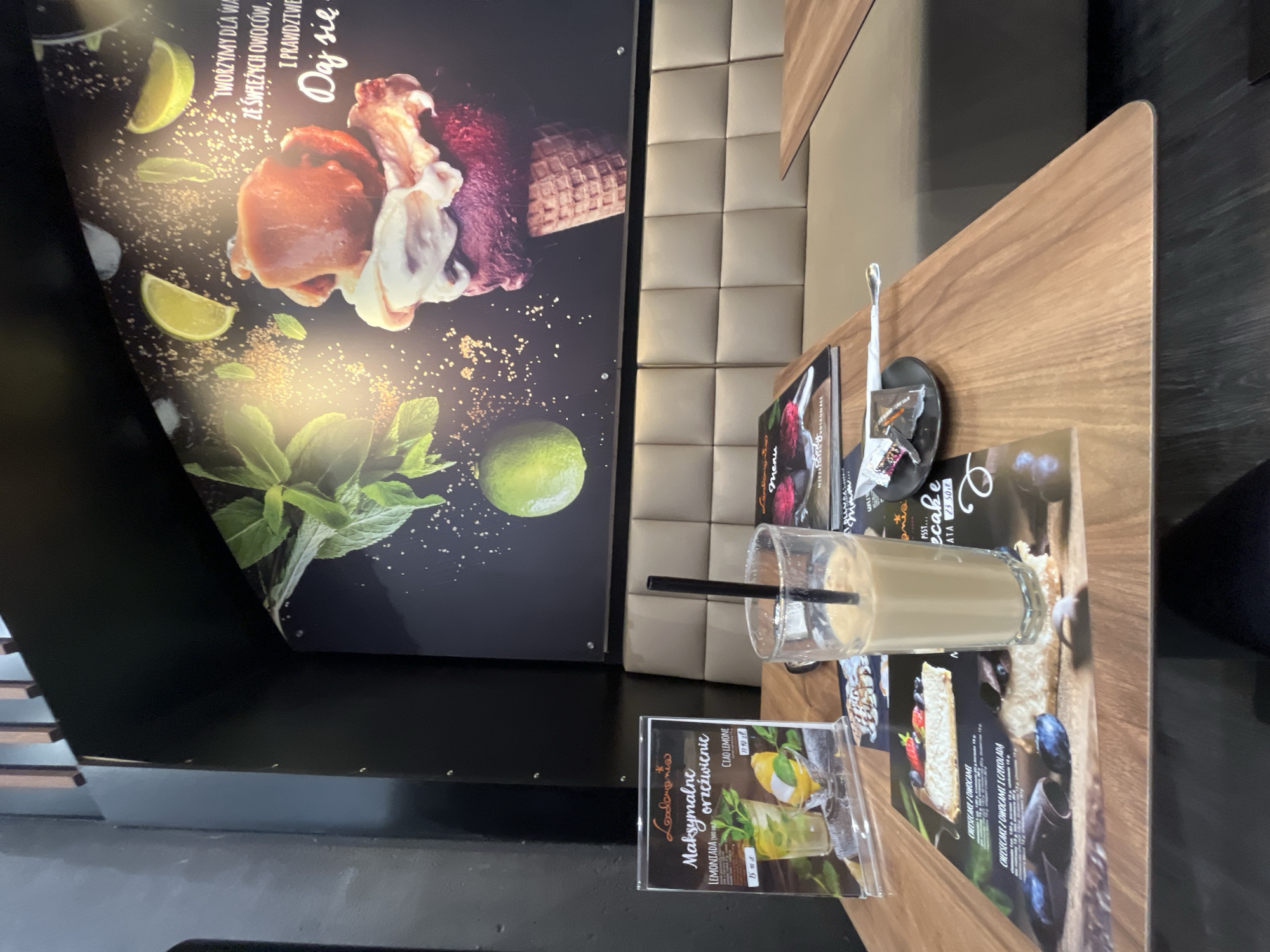 The image captures a close-up view of a wooden table in what appears to be a cafe. A tall glass filled with a light brown beverage, likely coffee or a milkshake, sits centrally on the table, complete with a black straw. To the left of the drink, a menu display showcases a picture of a lemonade with mint and lemon slices, and to the right, a smaller menu or placemat displays images of cheesecakes and other desserts. A small black saucer holding a napkin and what appears to be sugar packets sits near the edge of the table. In the background, a padded beige bench seating area is visible, and a large, dark wall mural features graphics of mint leaves, lime slices, and a layered ice cream cone. The overall ambiance suggests a modern and inviting cafe setting.