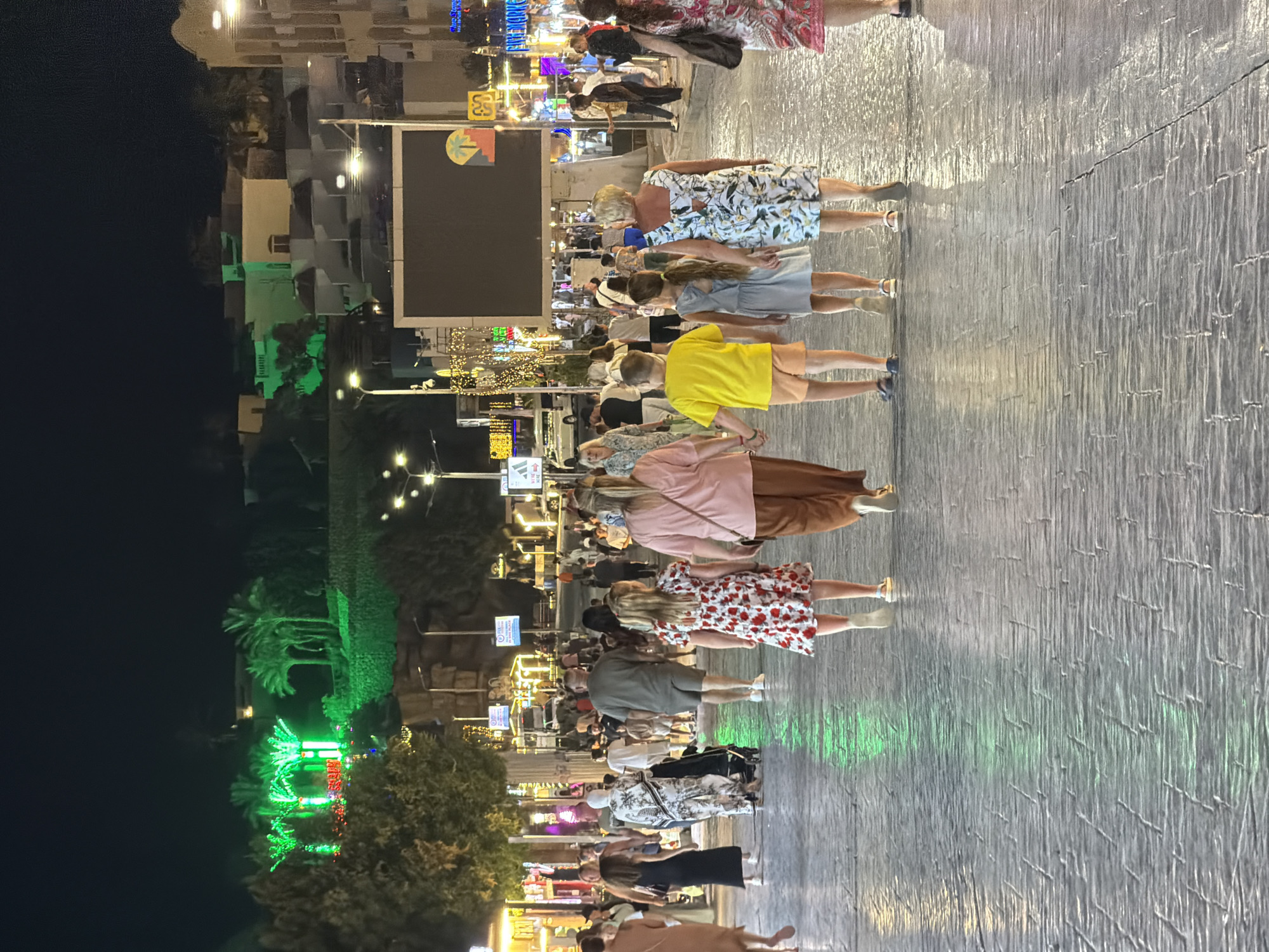 In Sharm El Sheikh, Egypt, at night, a paved promenade is bustling with people. The street is illuminated by ambient lighting from shops and buildings lining the walkway. In the background, illuminated palm trees and buildings are visible on a hillside, with one building facade lit in bright green. A large, dark screen is mounted on a building in the mid-ground. A group of people, including children, are walking away from the camera, holding hands. Other individuals and groups are also seen walking along the promenade. The ground is paved with textured stones that reflect the light.