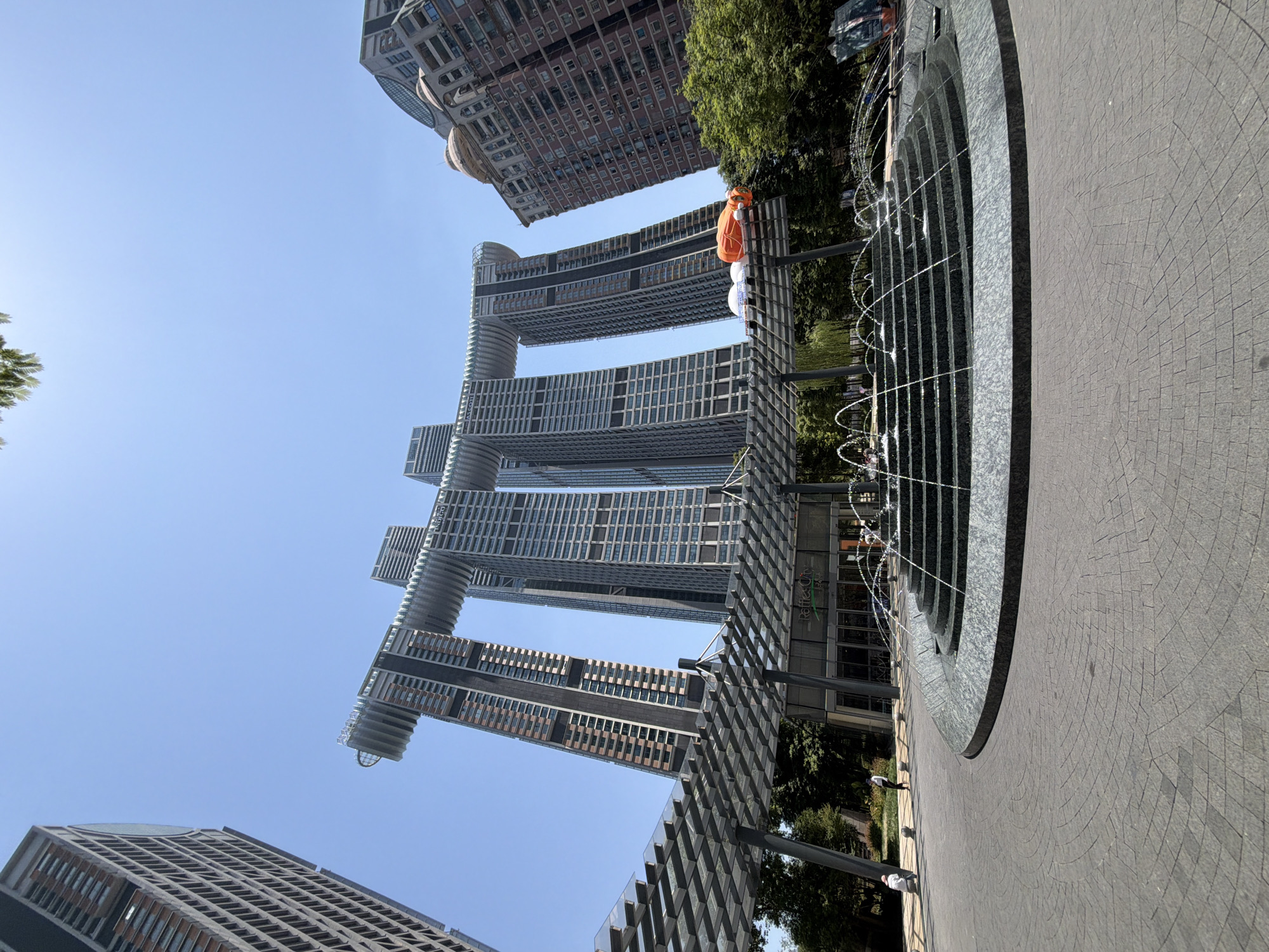 The image, taken from a low angle, captures the iconic Raffles City Chongqing in Yuzhong District, China, under a clear blue sky. The unique architectural design of the building, featuring four connected towers that resemble a ship, dominates the background. In the foreground, a tiered fountain with flowing water is set against a circular paved plaza. A modern-looking canopy structure with a transparent roof extends from the left of the frame towards the building. In front of the main building, a glass-fronted establishment with signage visible reads "Raffles City". To the right of the fountain, a large orange inflatable object resembling a goldfish is positioned on top of a white structure, partially obscured by the canopy. Several trees and greenery are visible behind the fountain and to the right of the building. Two individuals are visible walking on the paved area in the middle ground, and another person is seated on a bench to the left. The overall scene depicts a modern urban public space with distinctive architecture and public amenities.